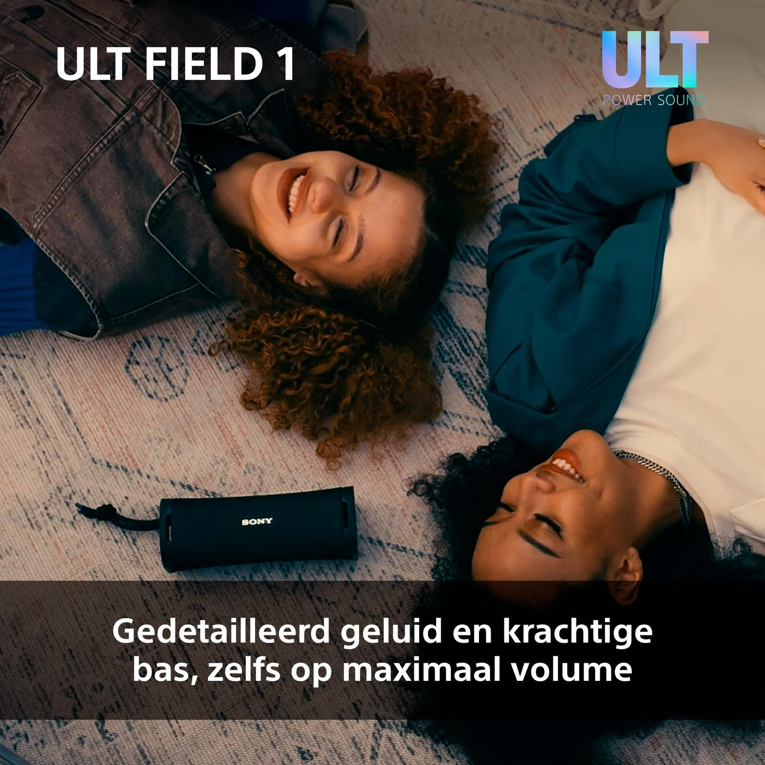Two smiling women lie on a patterned rug beside a black Sony portable speaker.