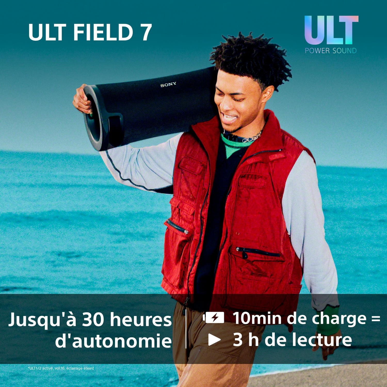 Un jeune homme souriant en gilet rouge porte une enceinte Sony noire sur son épaule près de la mer.