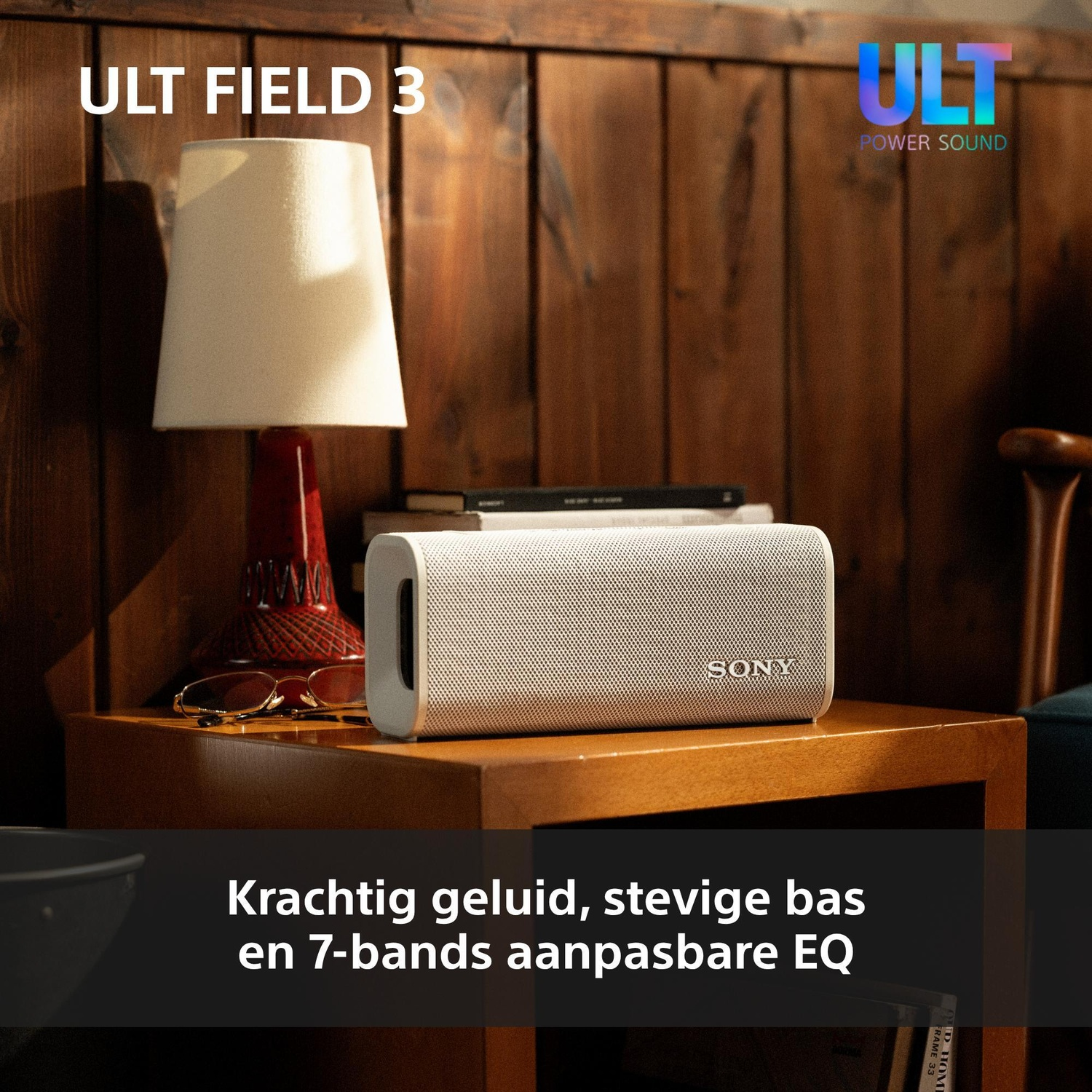 A white Sony portable speaker on a wooden nightstand beside a lamp, books, and glasses against a wood panel wall.