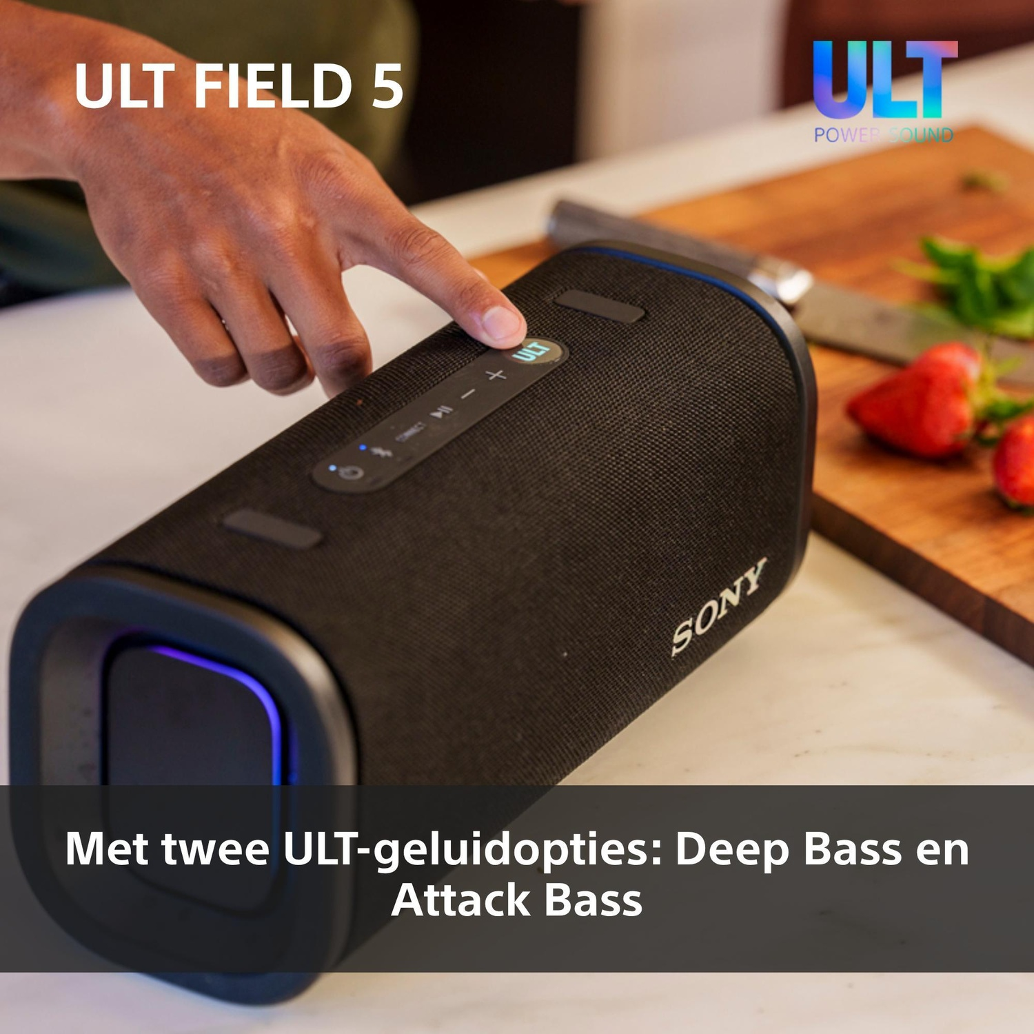 Hand pressing the ULT button on a portable Sony speaker in a kitchen with strawberries on a board.