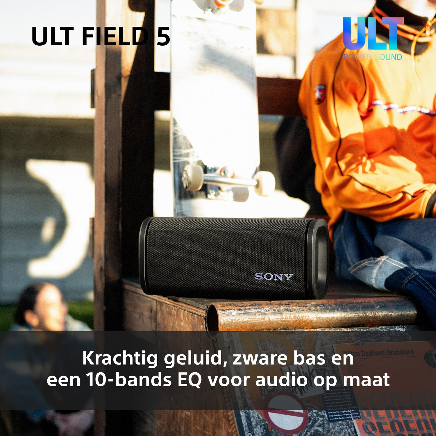 A black Sony portable speaker rests on a wooden surface outdoors, near a skateboard and a person in an orange jacket.