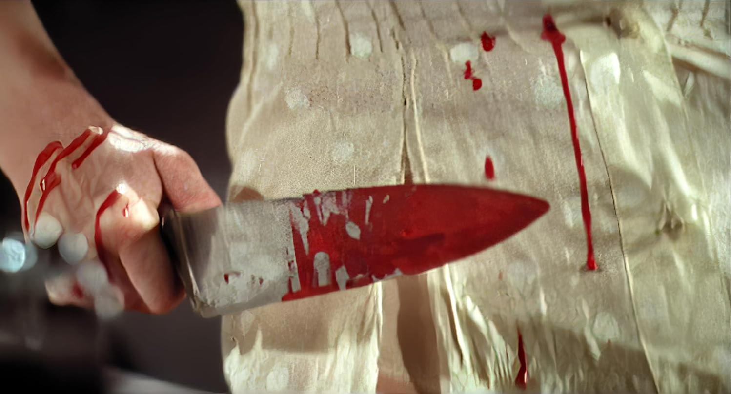Nahaufnahme einer Hand, die ein blutiges Messer vor einem verschmutzten Stoff hält.