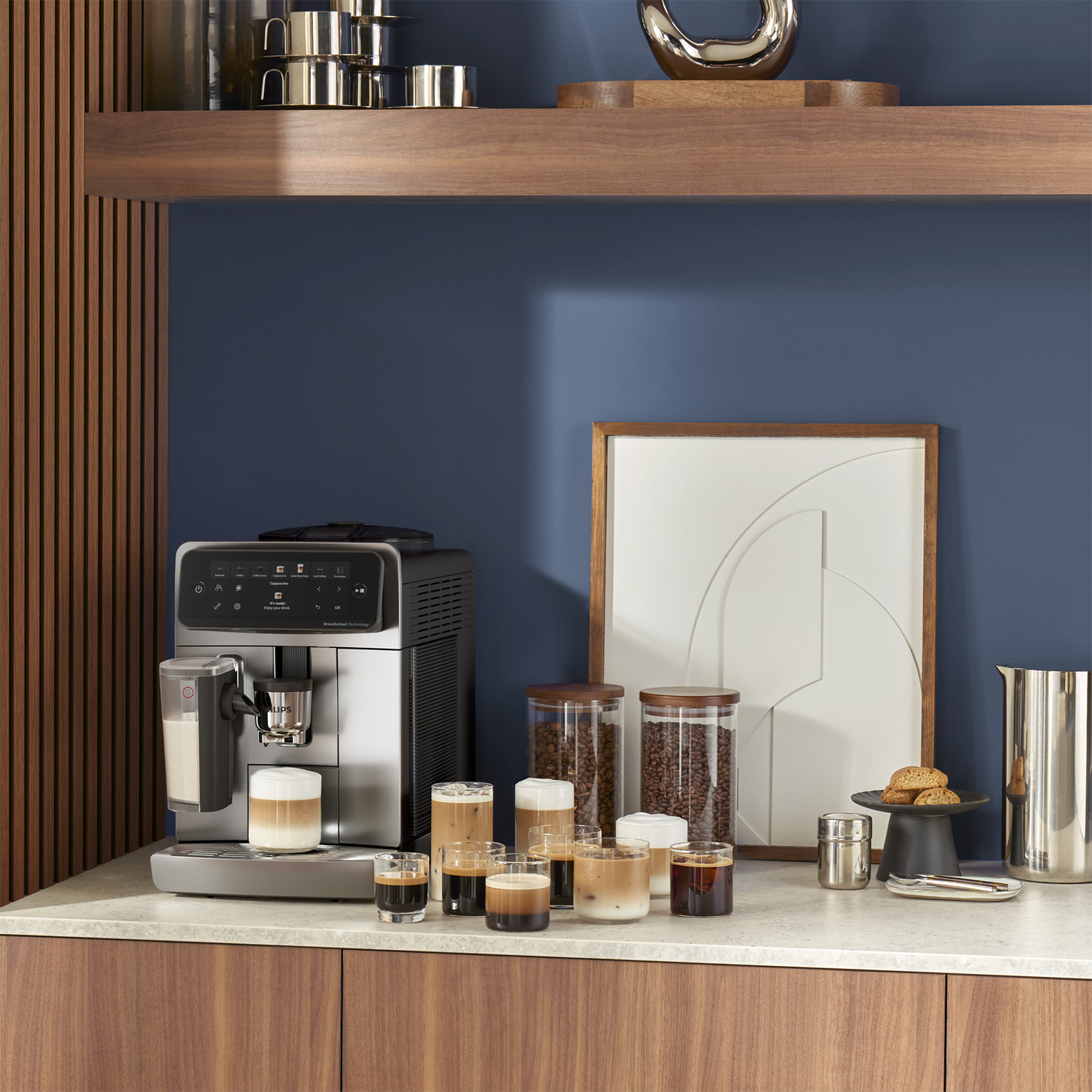 Modern coffee machine on a marble countertop with coffee glasses, bean jars, and framed art against a blue wall.