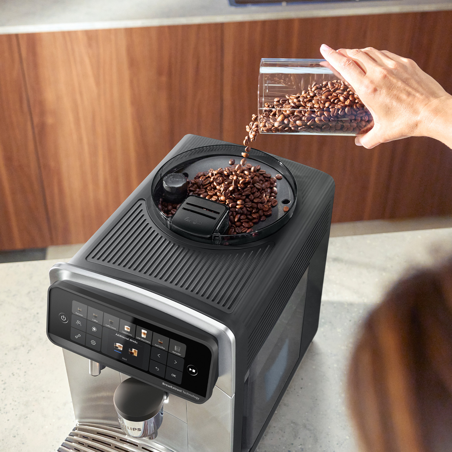 A hand pours coffee beans from a container into the top hopper of a modern black and silver espresso machine.