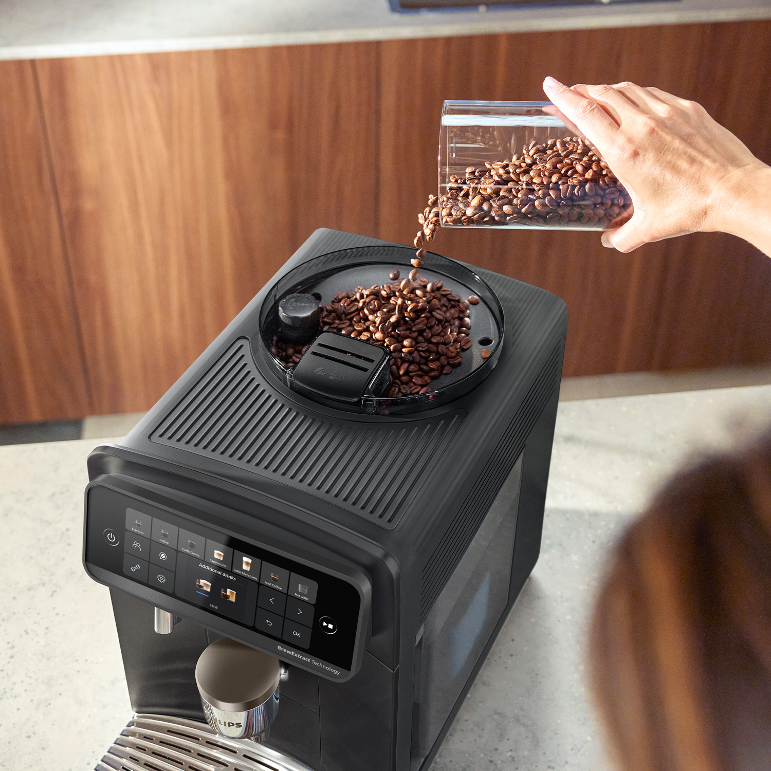 A hand pours coffee beans from a container into the grinder hopper of a black coffee machine.