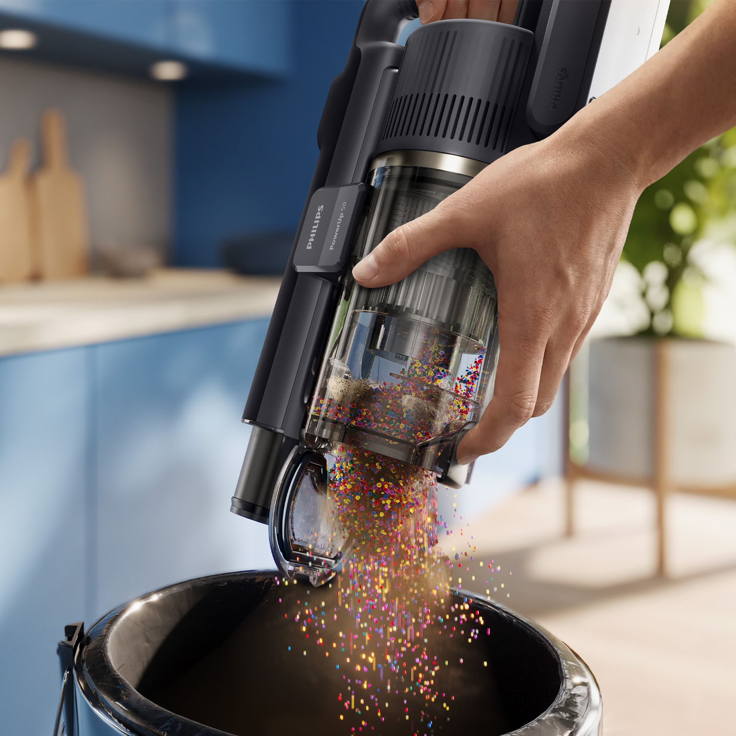 A hand empties a black cordless vacuum's dust bin, spilling colorful beads into a black bucket in a kitchen.
