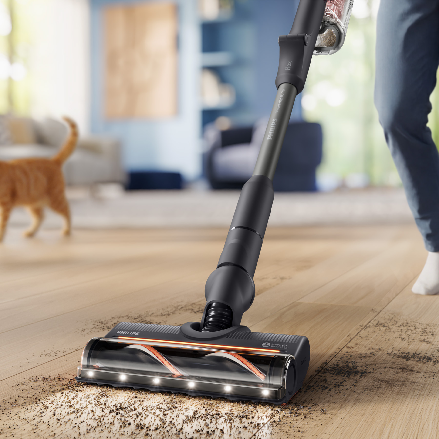 A cordless vacuum cleans debris from a wooden floor, with a cat visible in the blurred background.