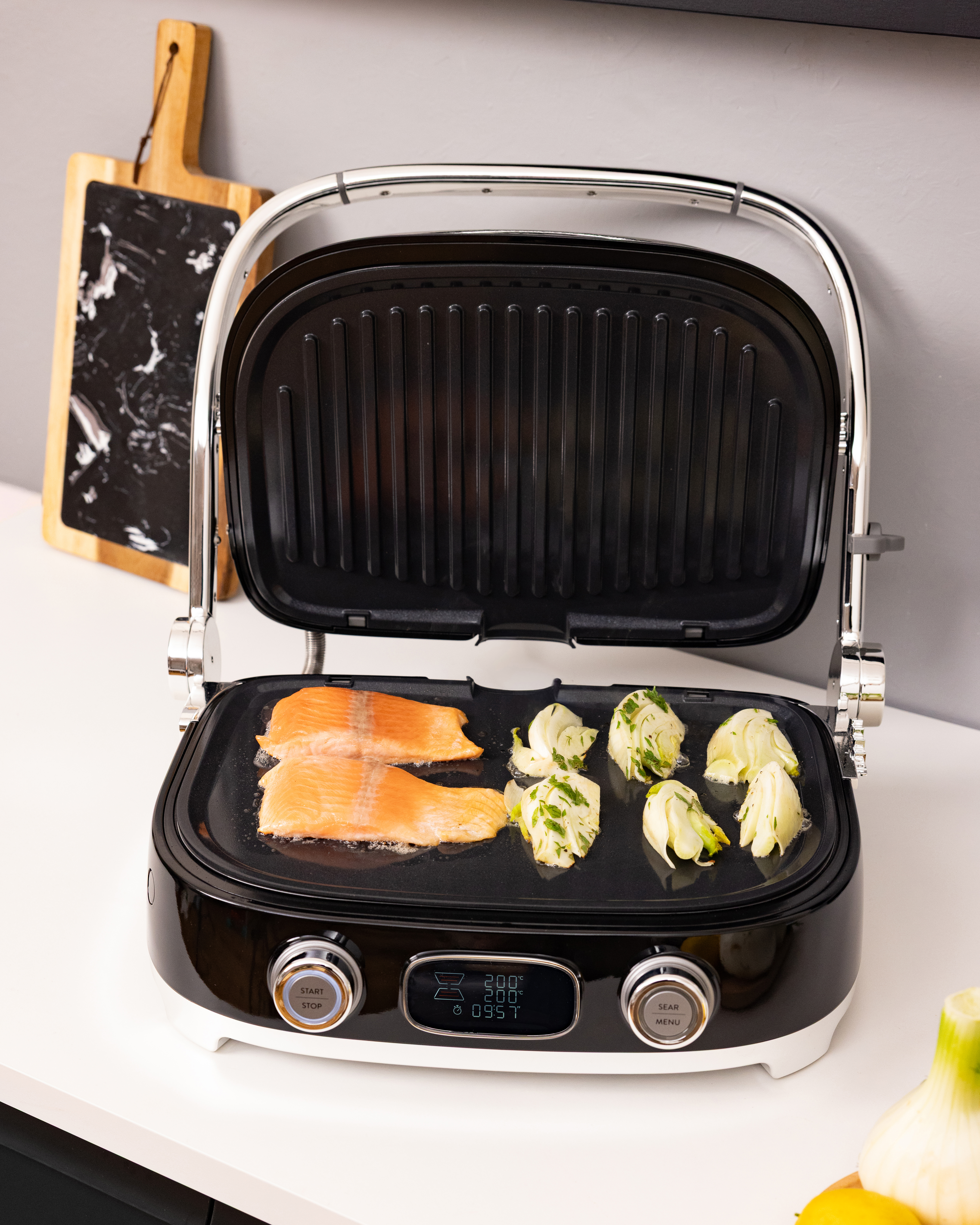 Lachsfilet und Fenchel werden in einem schwarzen Tischgrill auf einer weißen Theke zubereitet.