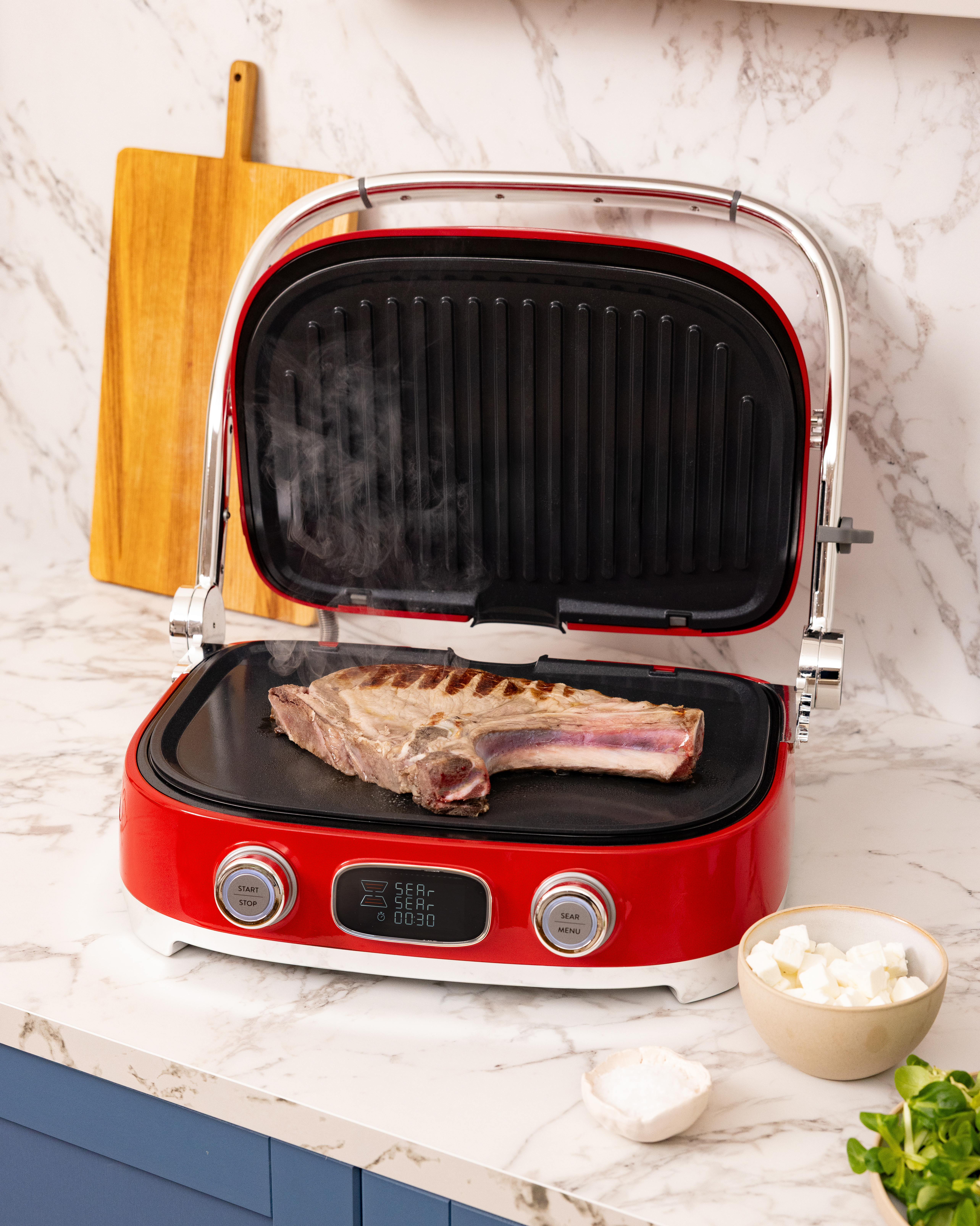 Ein rohes Steak mit Knochen gart auf einem dampfenden roten Elektrogrill auf einer Marmorarbeitsplatte.