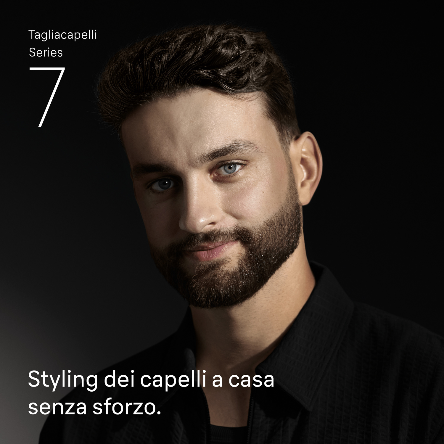 Primo piano di un uomo curato con barba e camicia scura su sfondo nero.