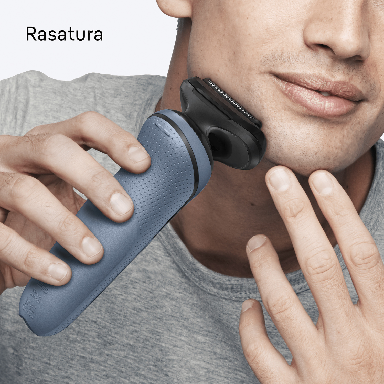 Un uomo con una camicia grigia si rade il viso con un rasoio blu e nero.