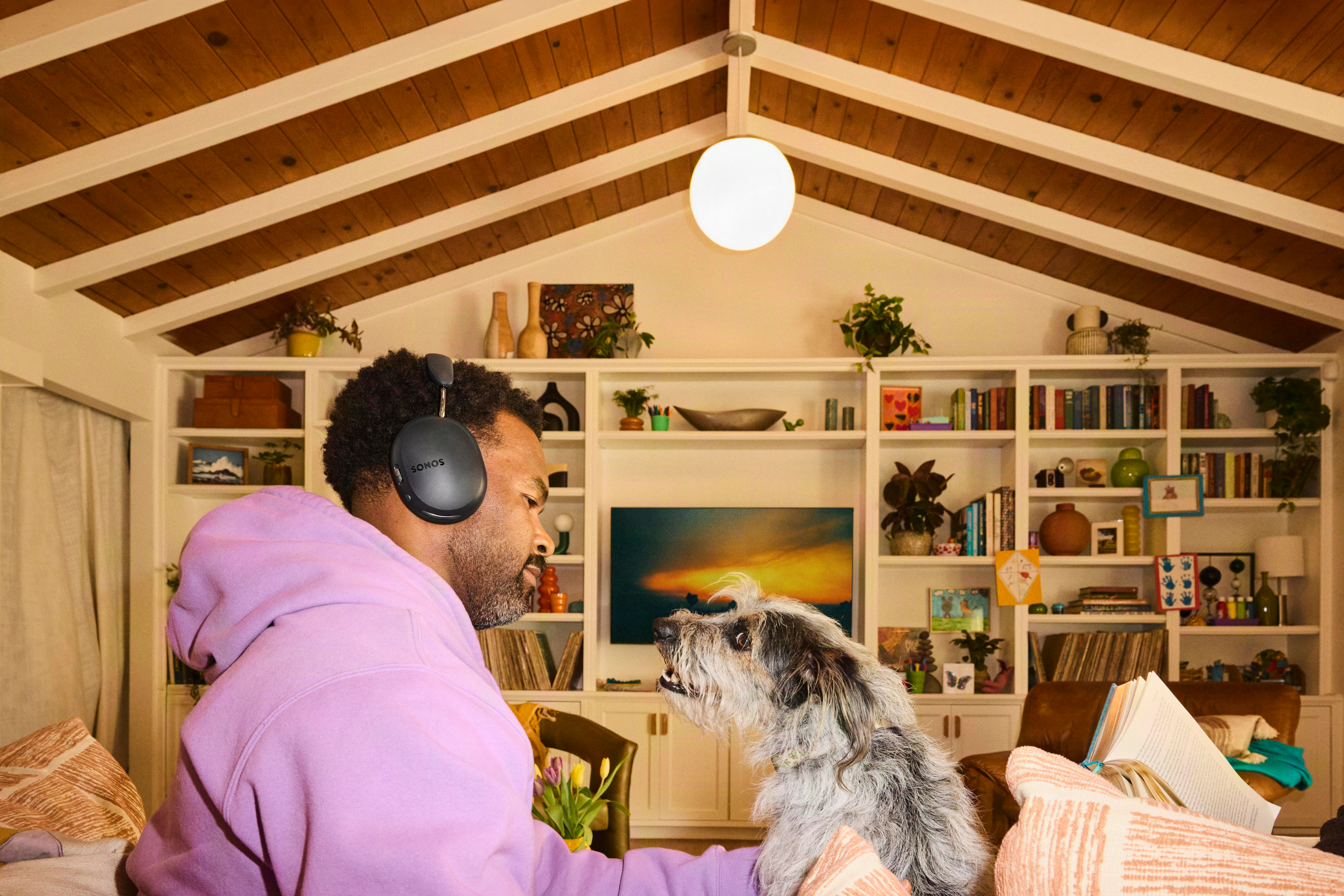 Ein Mann mit lila Kapuzenpulli und Kopfhörer schaut in einem Wohnzimmer mit Bücherregalen einen zotteligen Hund an.