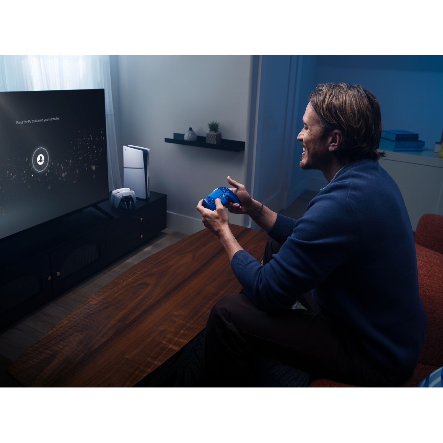A man sits in a living room holding a blue controller while playing video games on a large TV.