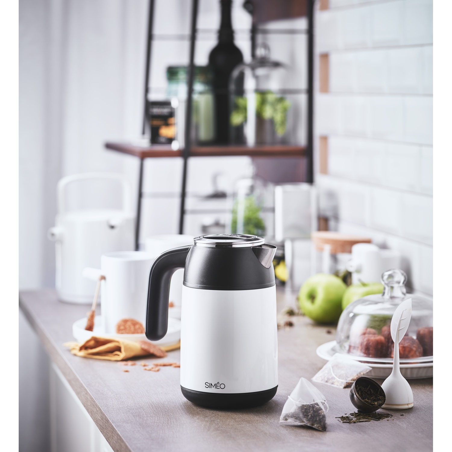 A white and black Simeo kettle on a kitchen counter with tea bags, green apples, and dishware in the background.