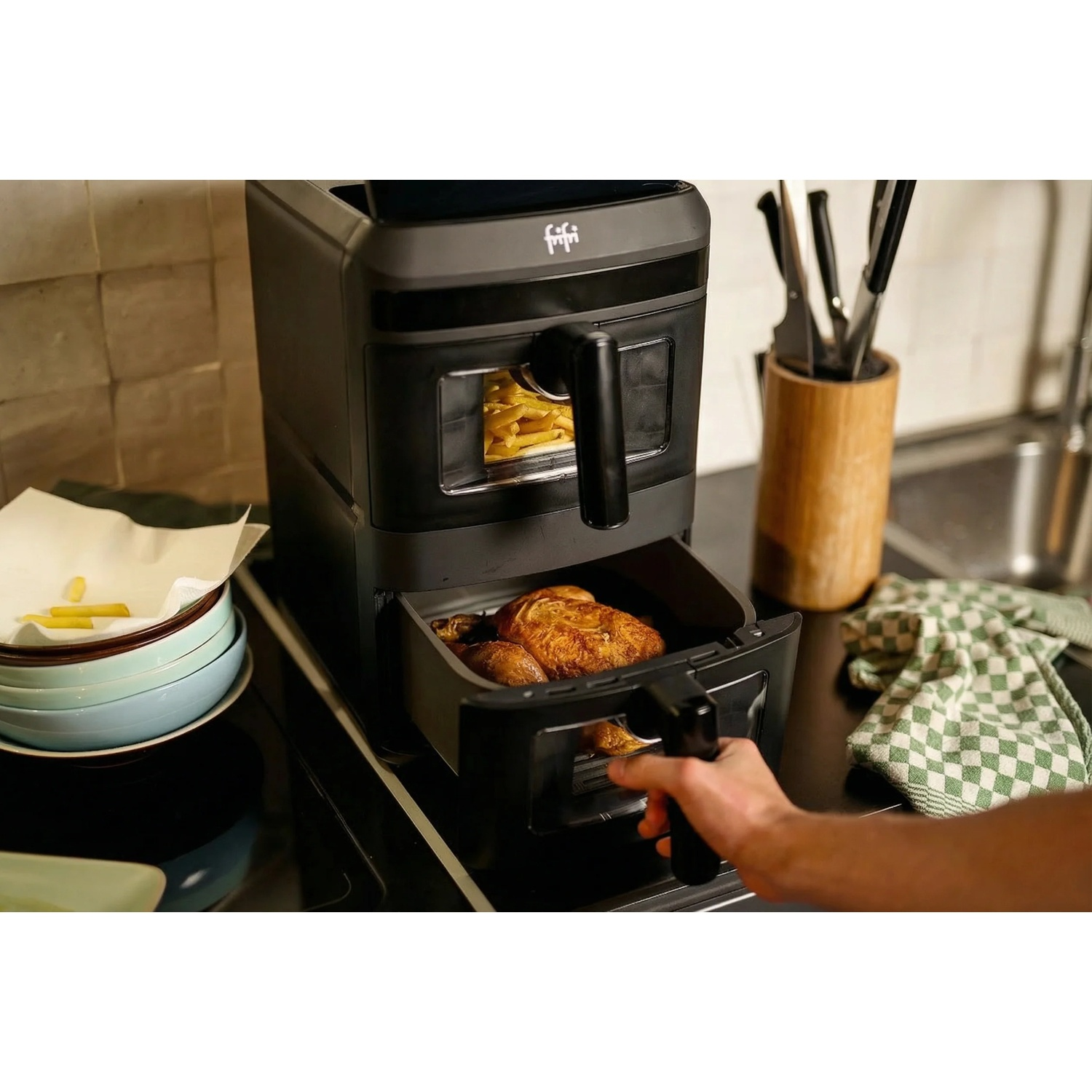 A hand opens the bottom drawer of a black air fryer, revealing a whole chicken roasting inside.