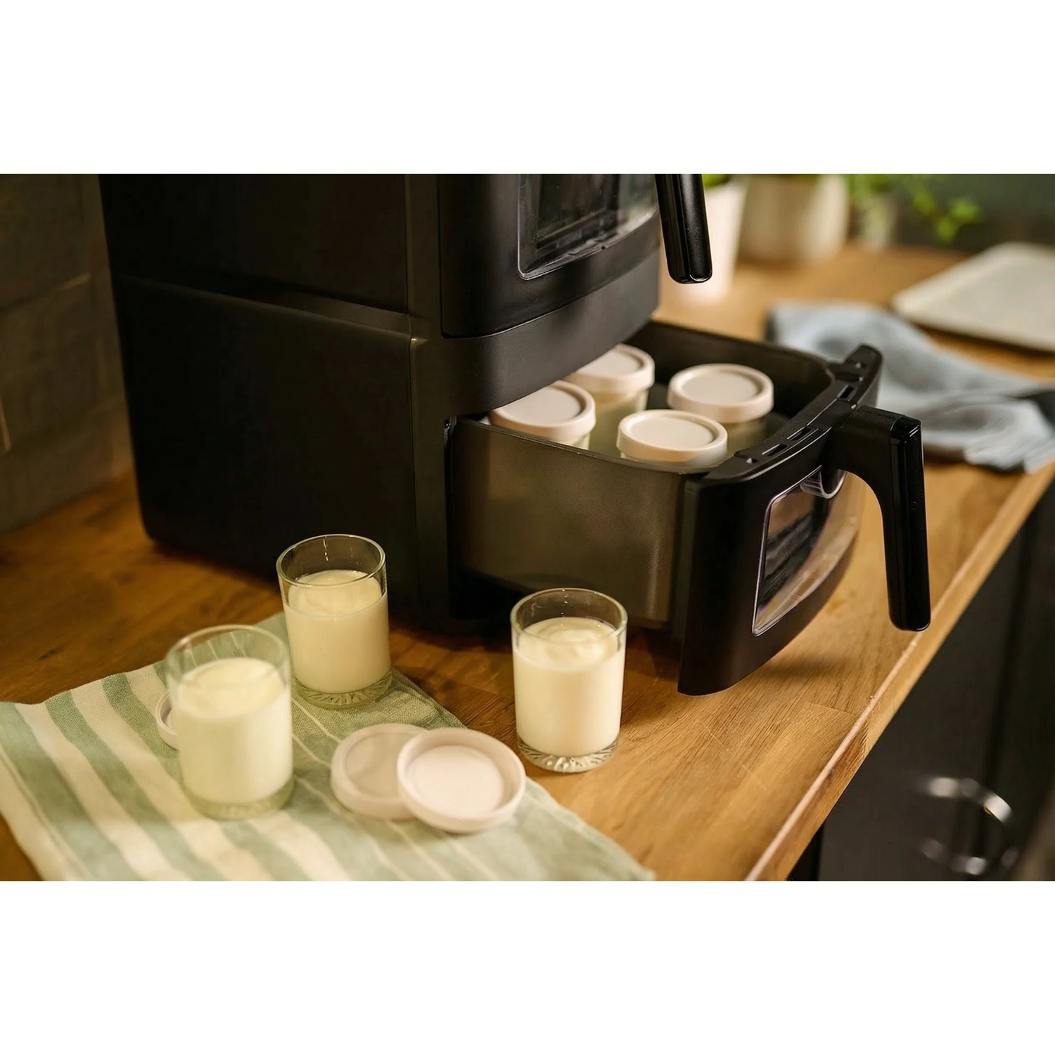 An air fryer on a wooden countertop with its drawer open, revealing jars of yogurt, with more jars placed outside.