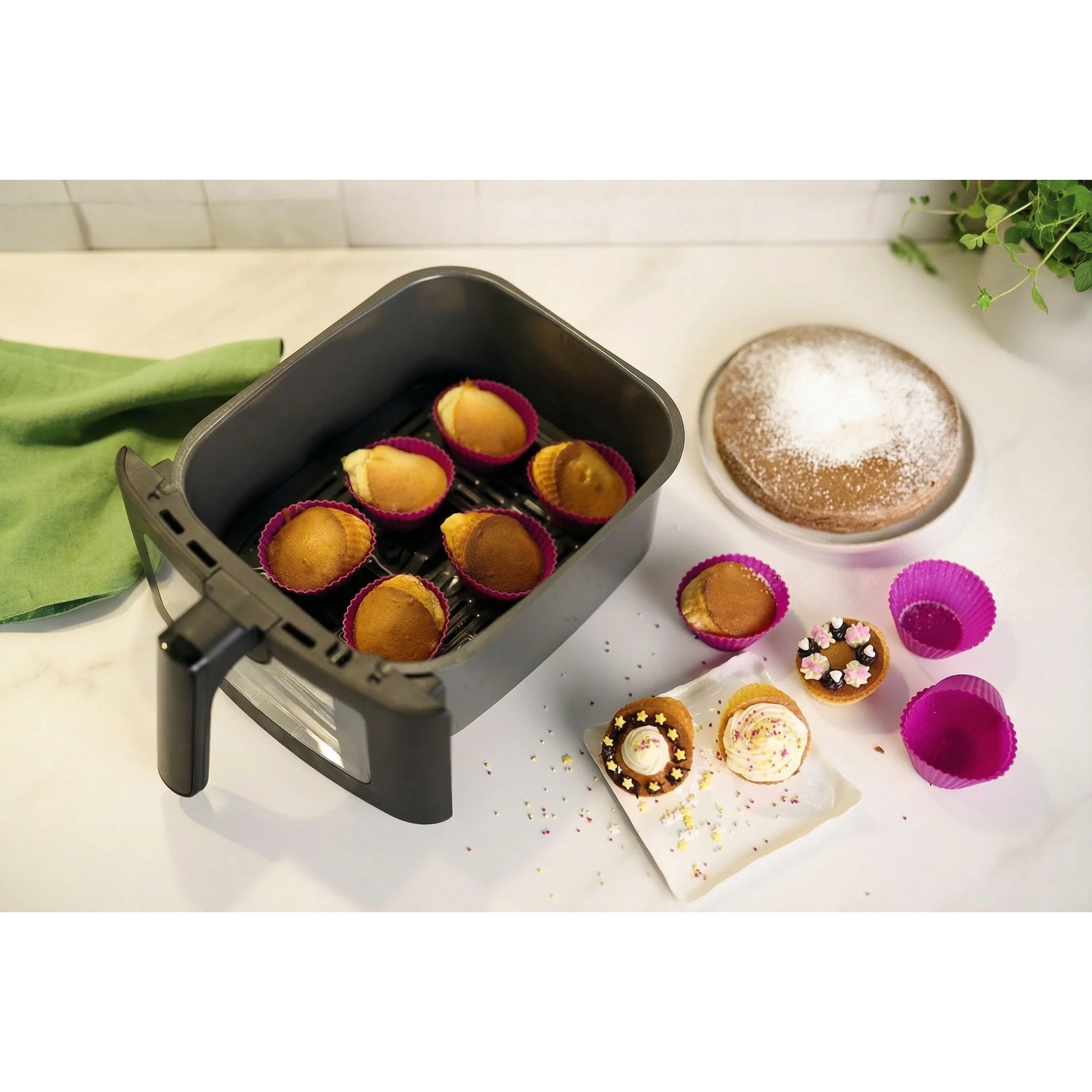 Muffins in an air fryer basket, a round cake with powdered sugar, and decorated cupcakes on a light countertop.