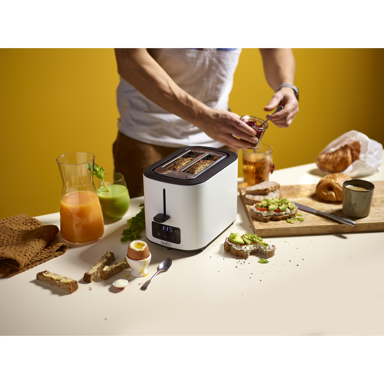 A person preparing breakfast next to a white toaster, juice, avocado toast, and croissants on a table.