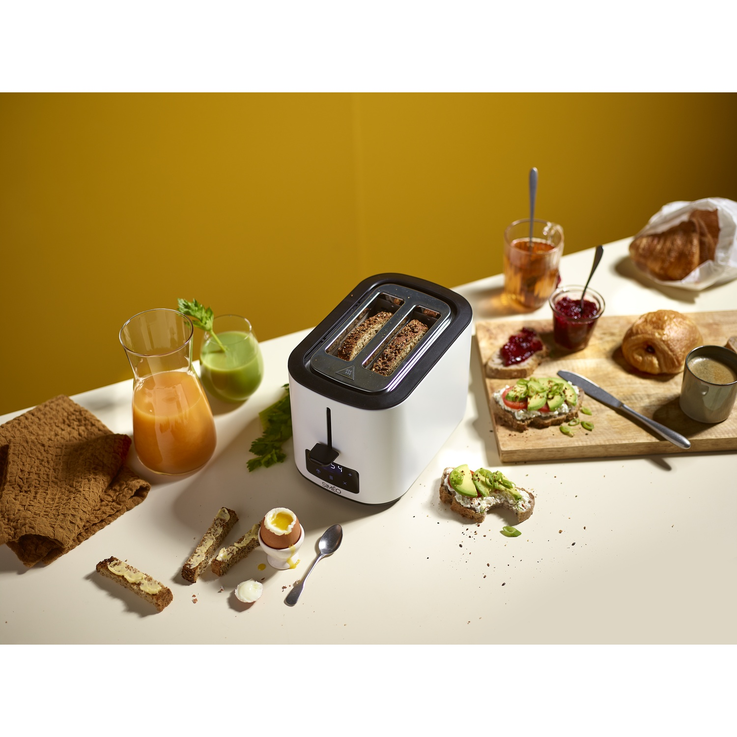 A white toaster on a breakfast table with bread, egg, juice, and spreads against a yellow wall.