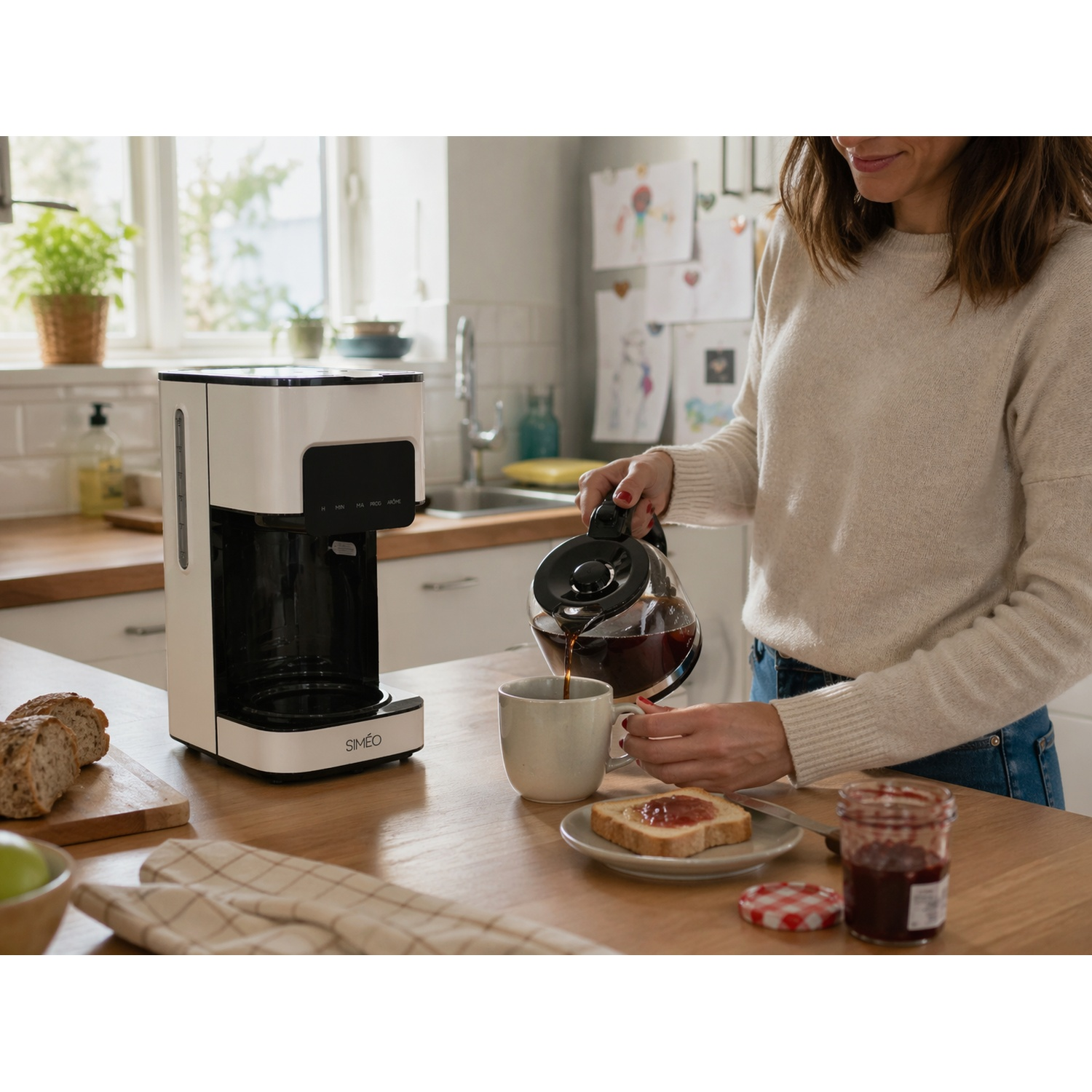 A woman pours coffee into a cup in a kitchen next to a coffee maker, bread, and jam.