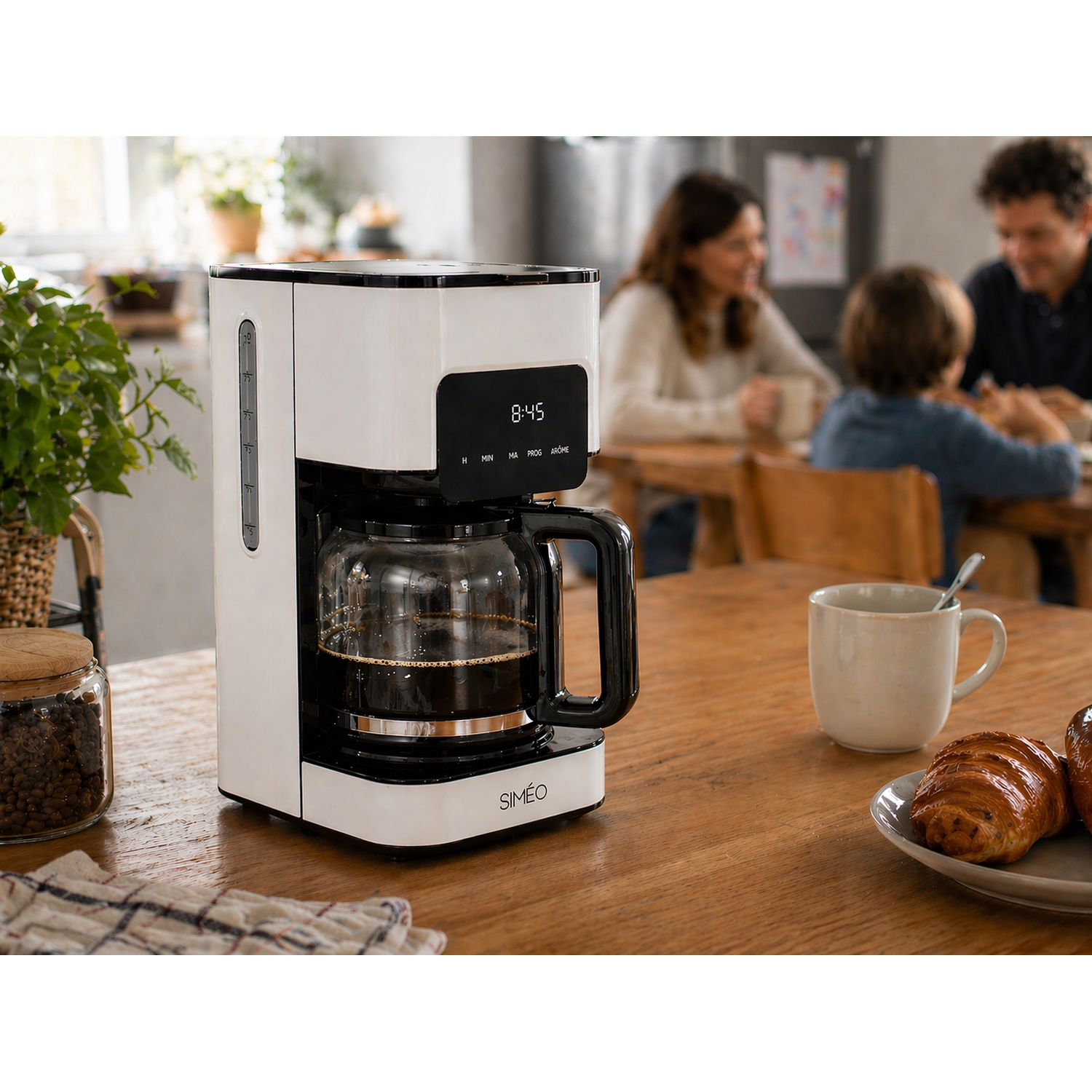 A white and black drip coffee maker on a wooden kitchen table, with a family having breakfast in the blurred background.