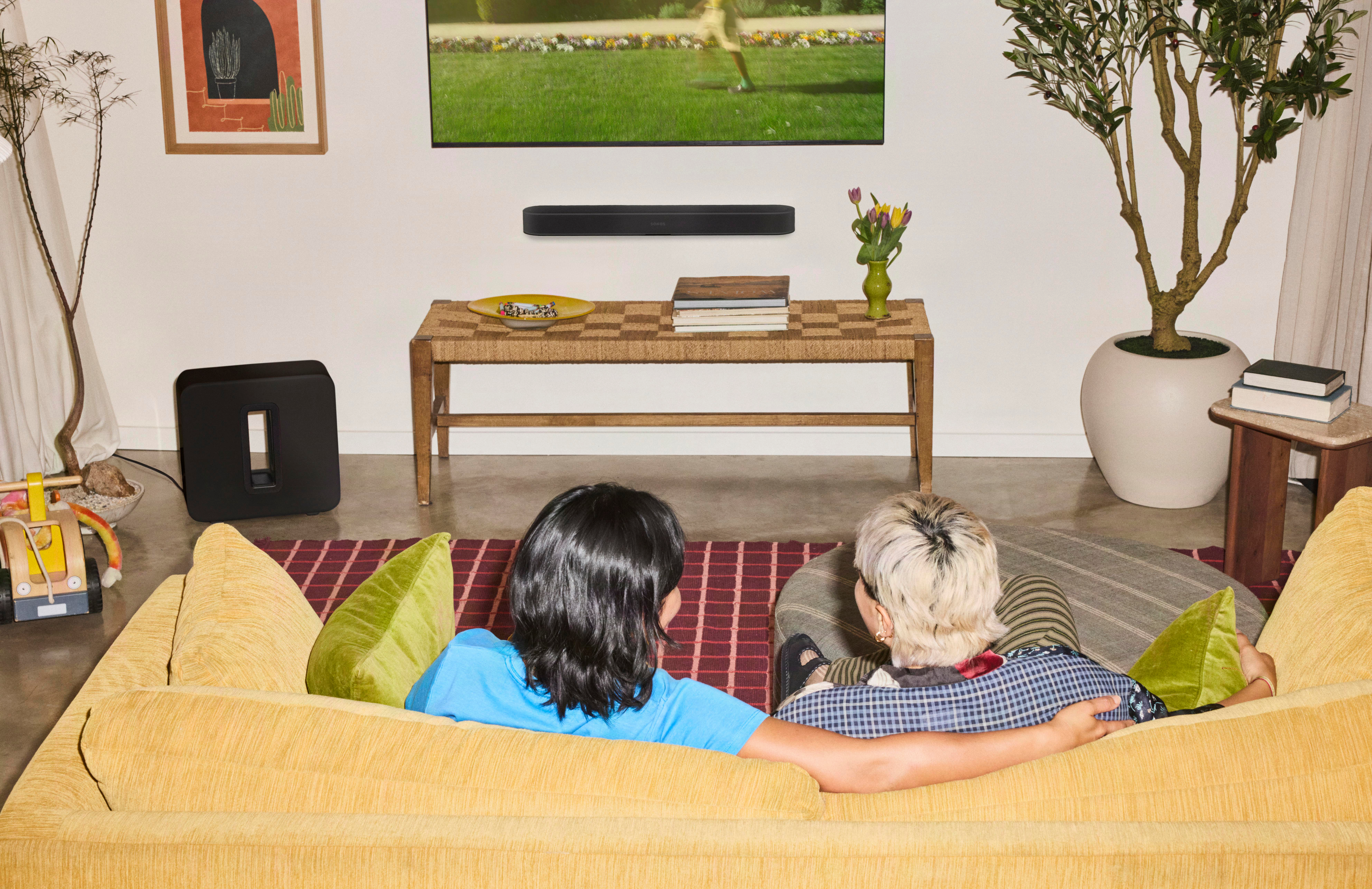 Zwei Personen sitzen auf einem gelben Sofa und schauen fern im Wohnzimmer.