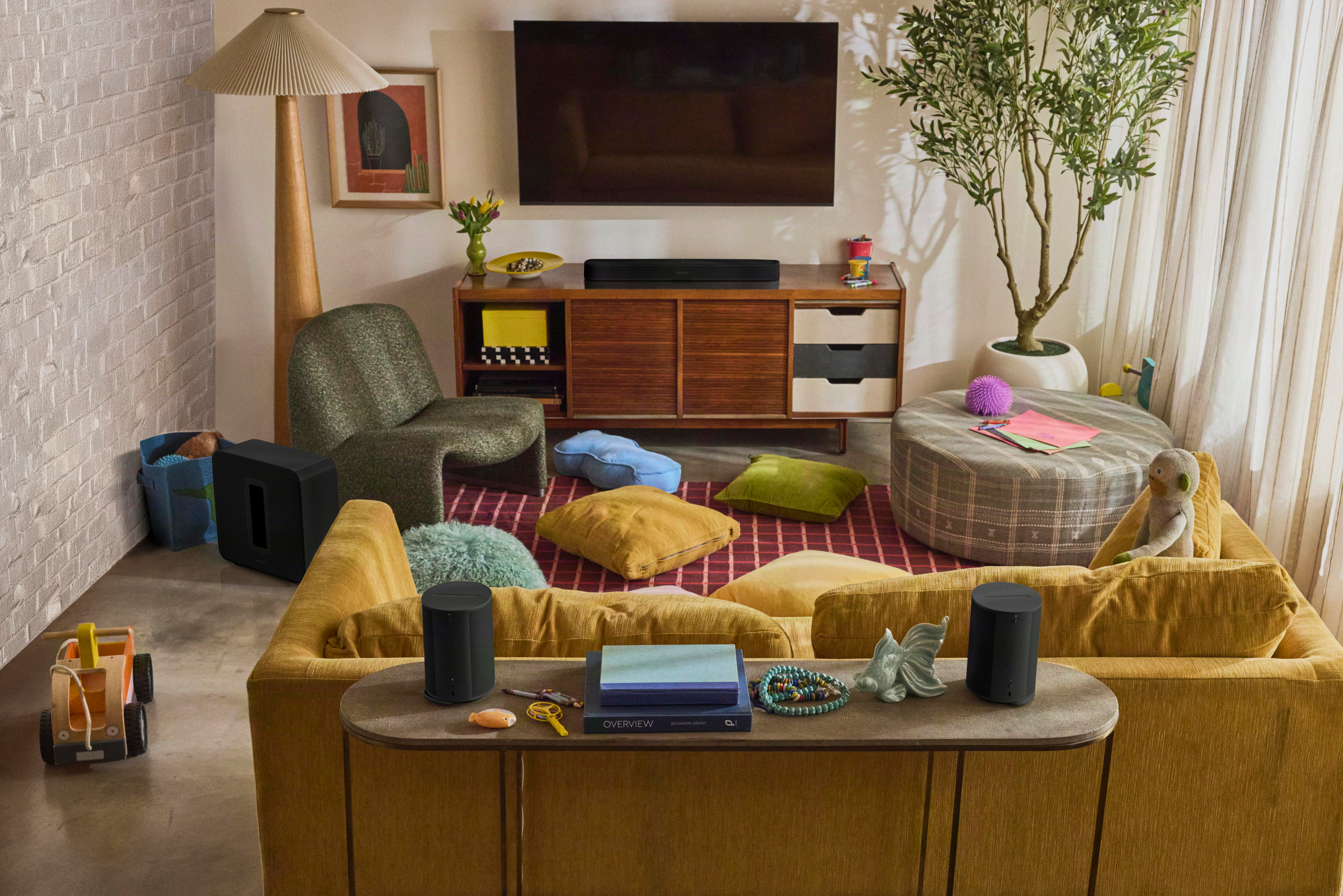 Wohnzimmer mit gelbem Sofa, TV, runder Bank, grünem Sessel, Holzkonsole, Pflanze und Stehlampe.