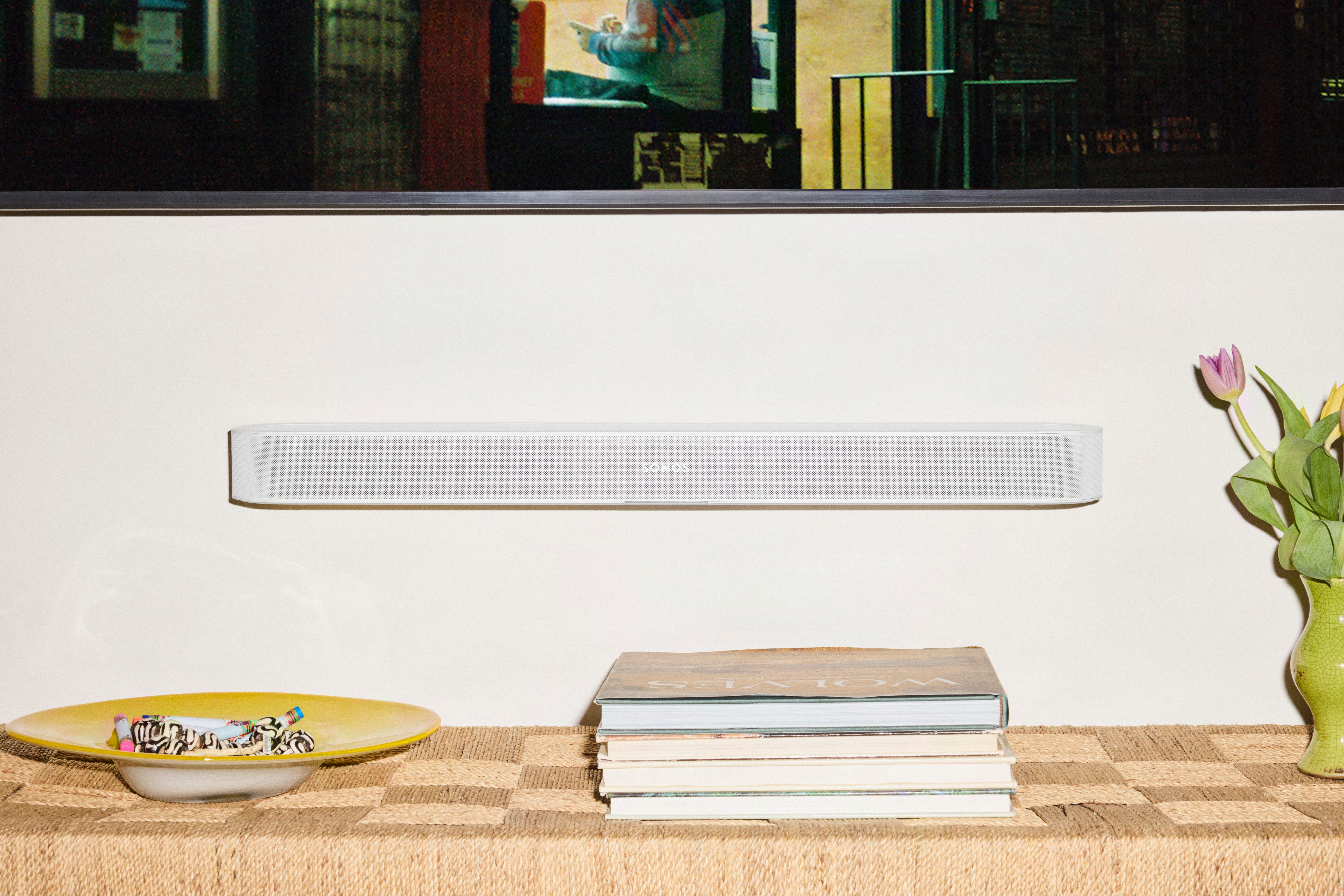 Eine weiße Soundbar hängt an einer Wand über einer Kommode mit Büchern, einer Schale und einer Vase mit Blumen.