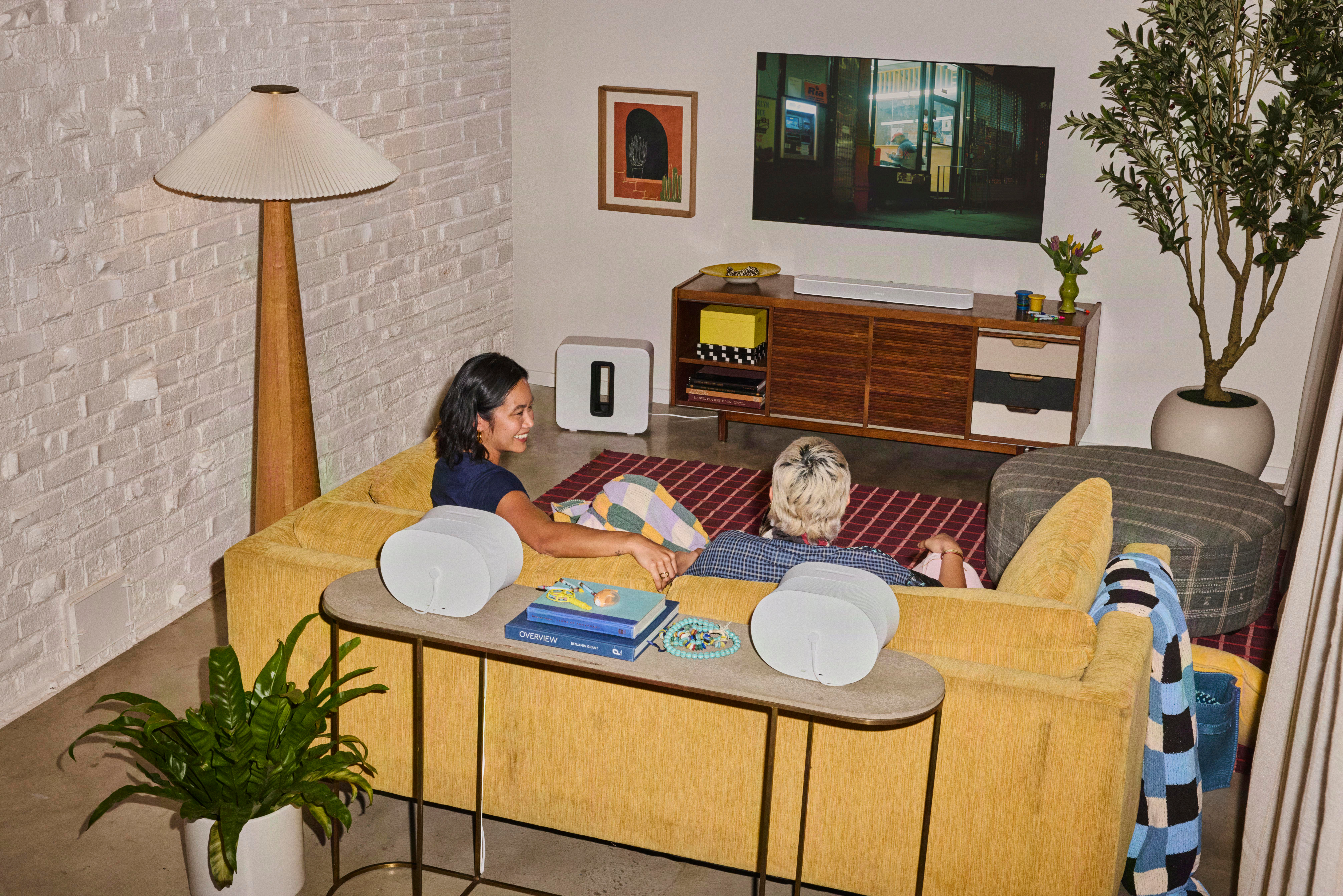 Zwei Personen sitzen auf einem gelben Sofa in einem modernen Wohnzimmer mit Fernseher und Pflanzen.