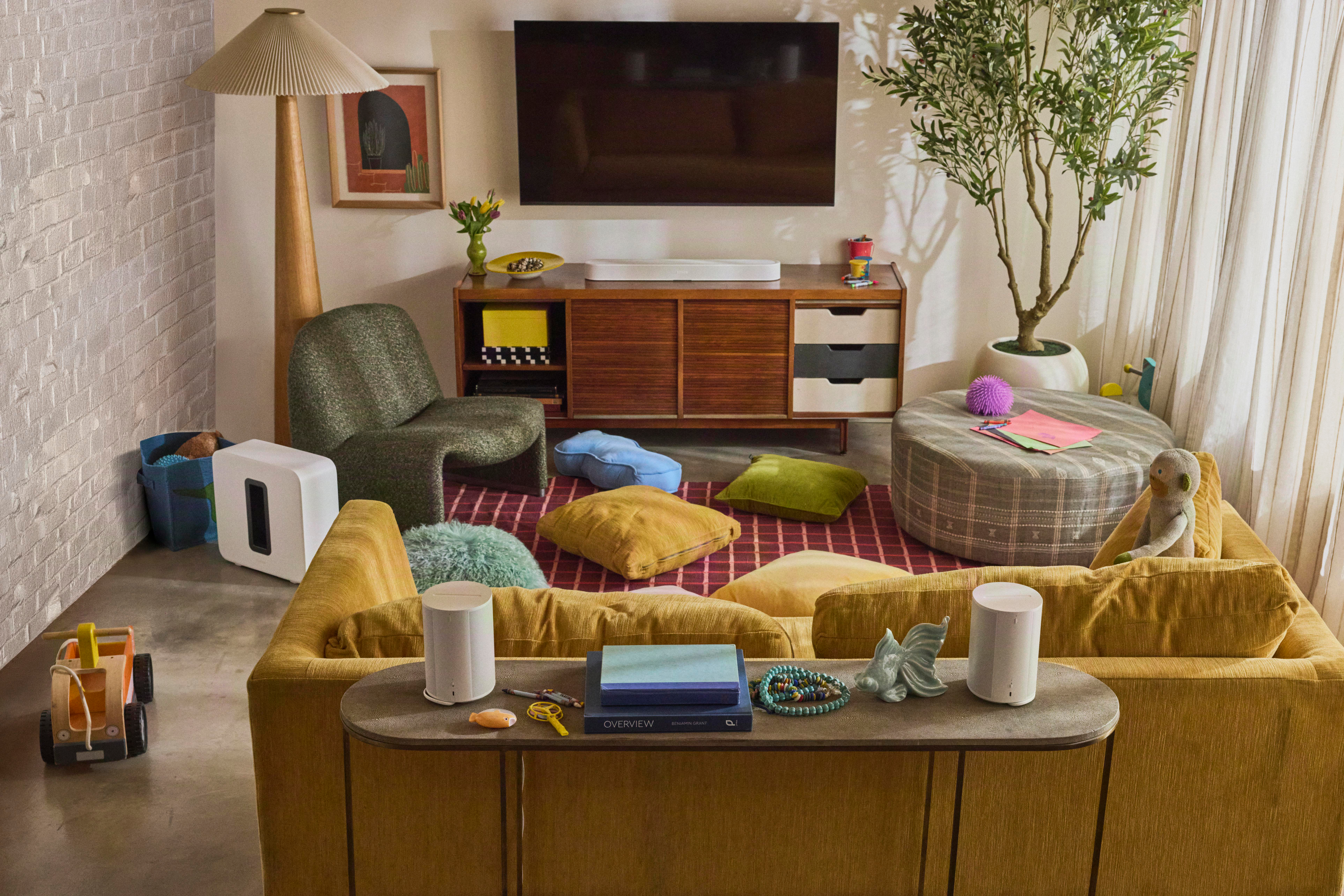 Gemütliches Wohnzimmer mit gelbem Sofa, TV, Holzkonsole, Teppich, Kissen, Pflanzen und Stehlampe.