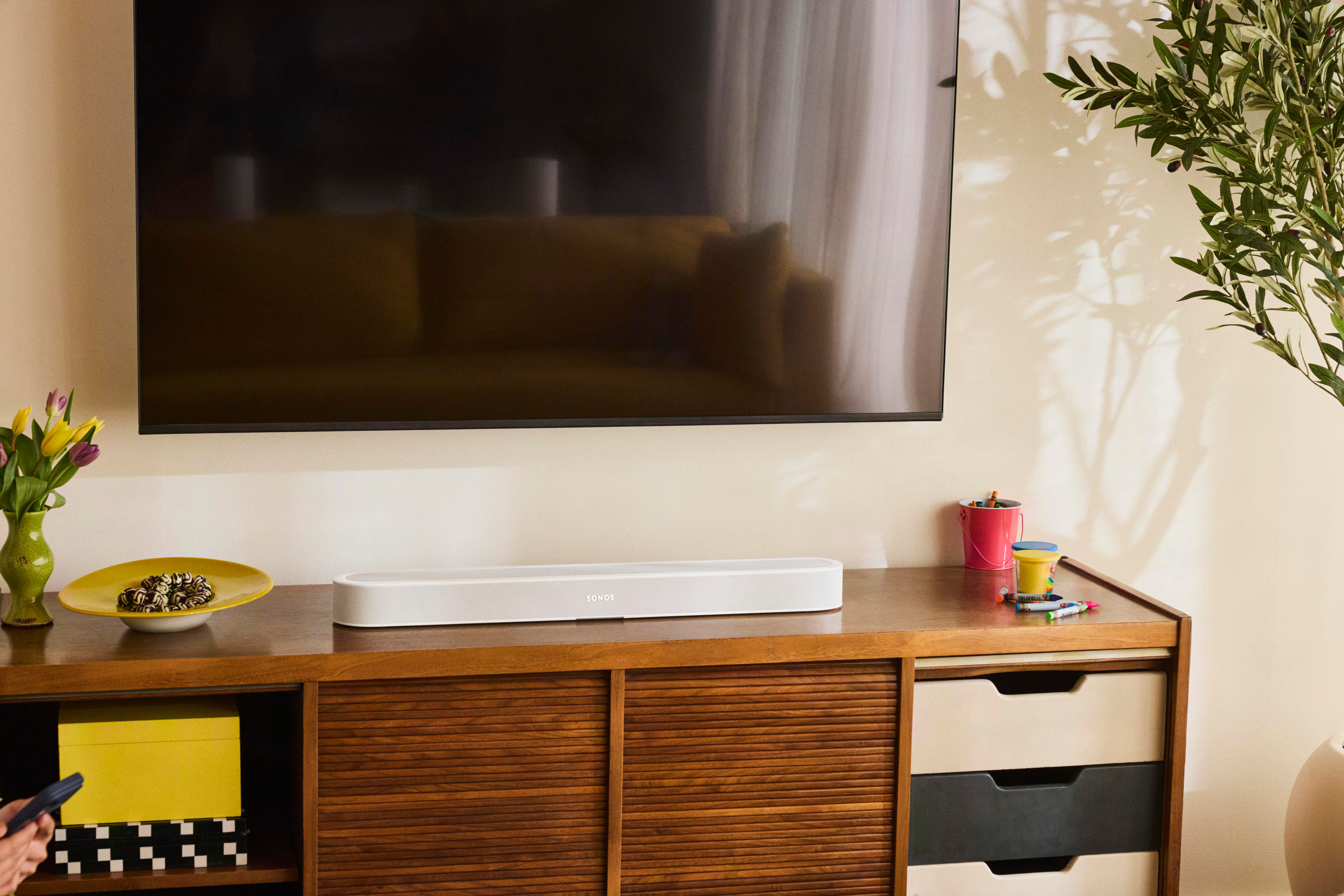 Eine Soundbar auf einer Holzkommode unter einem Wandfernseher, mit Vasen, Blumen und Schatten im Hintergrund.