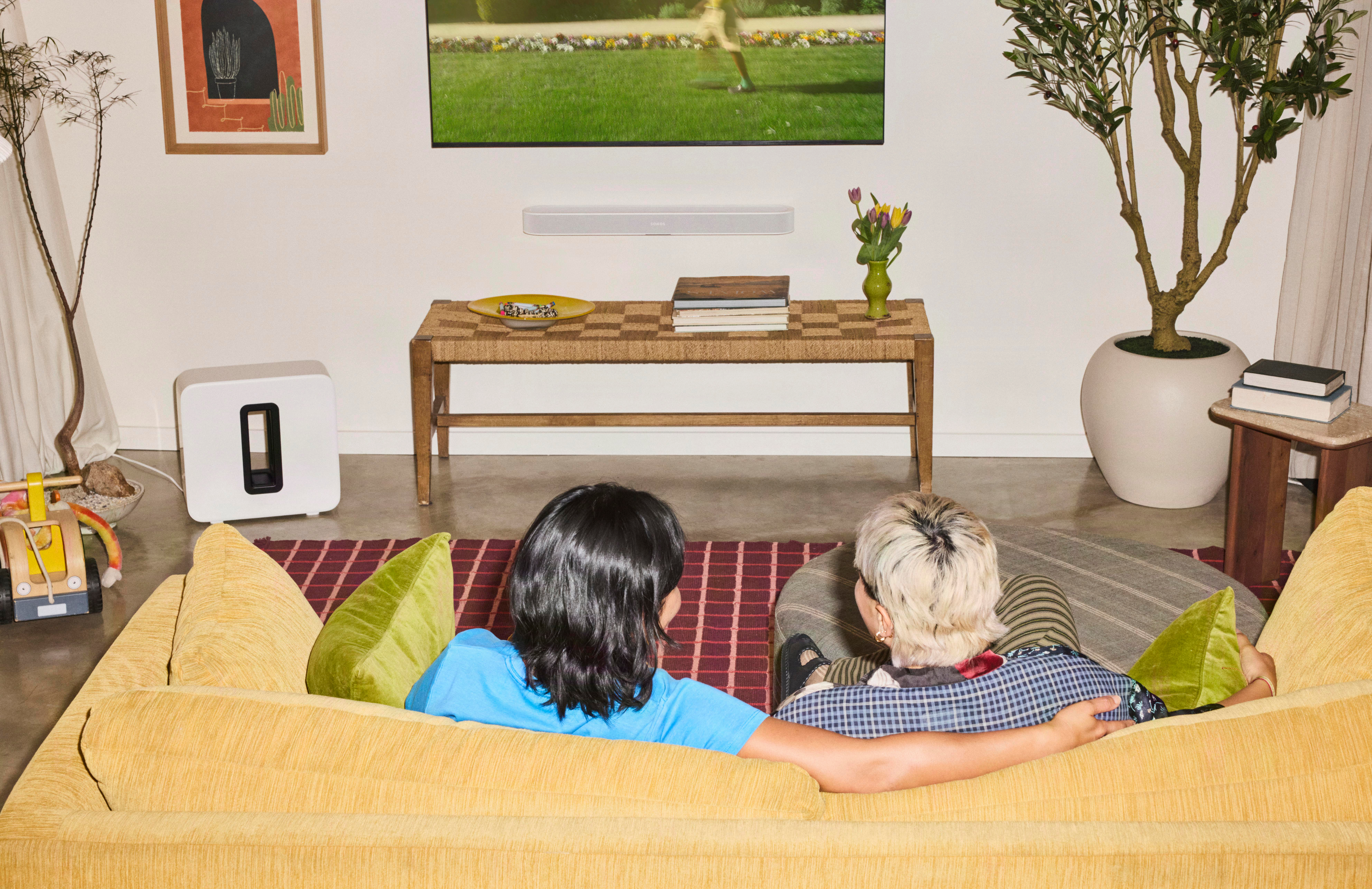 Zwei Personen sitzen auf einem gelben Sofa in einem hellen Wohnzimmer und schauen fern.