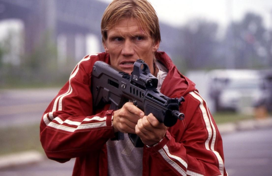 Ein blondhaariger Mann in einer roten Jacke zielt mit einem Sturmgewehr im Freien.