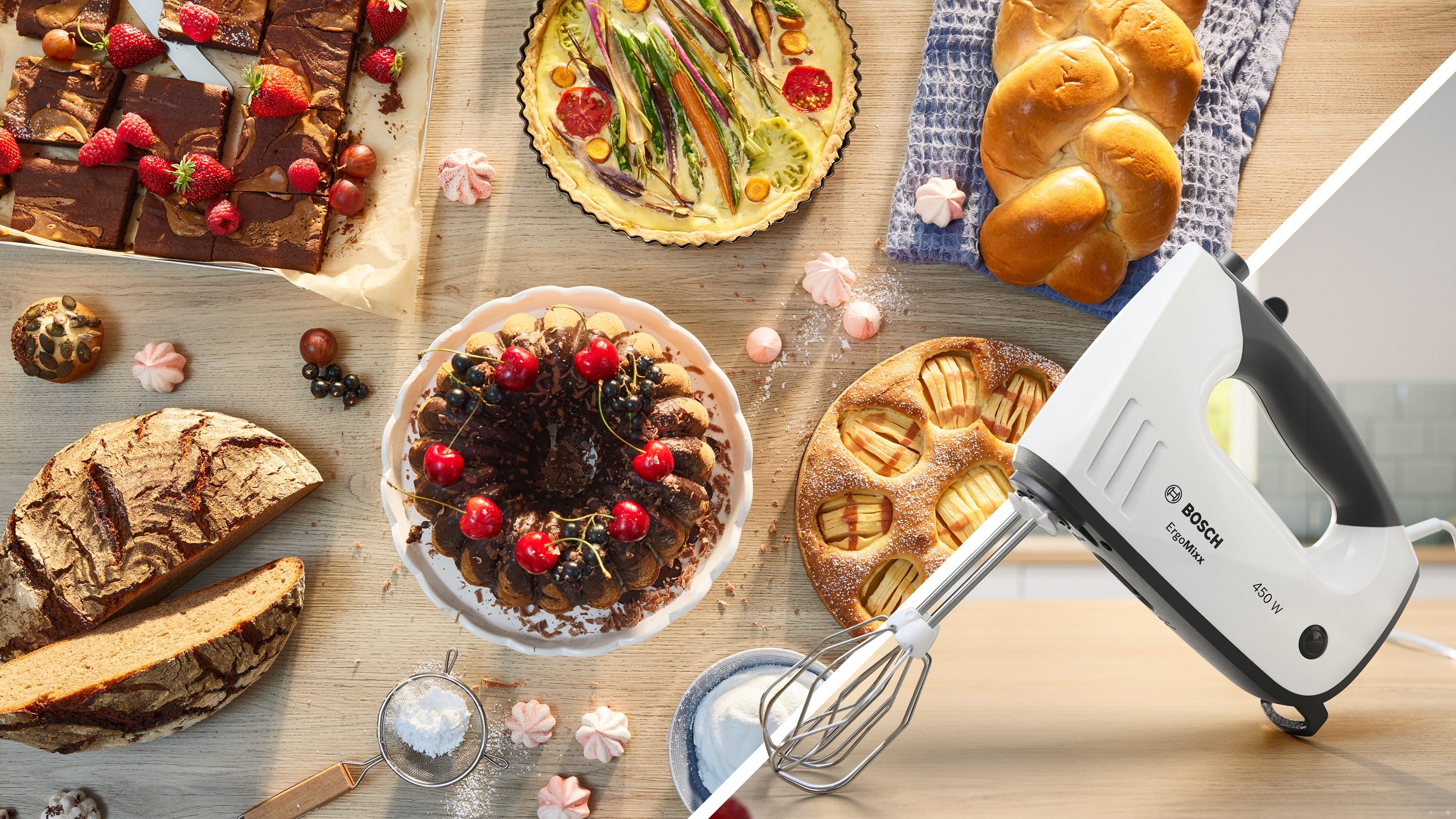 Ein weißer Bosch-Handmixer liegt auf einem Holztisch mit Kuchen, Brot und Beeren.