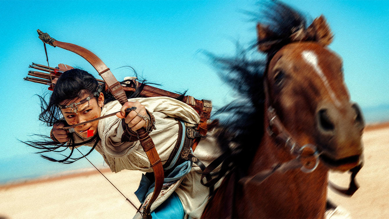 Eine Reiterin in traditioneller Kleidung spannt im Galopp einen Bogen.