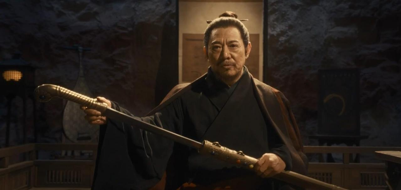 Ein asiatischer Mann in traditioneller Kleidung hält ein goldenes Schwert in einem Raum mit Steinwänden.