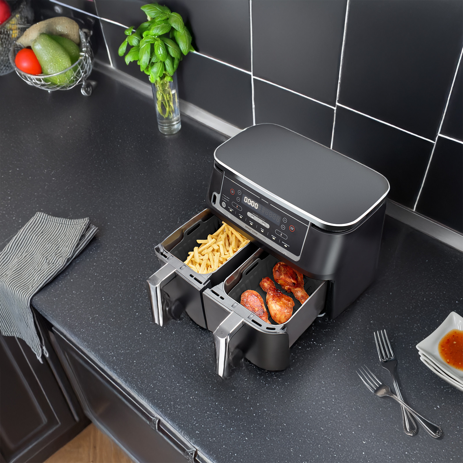 Eine schwarze Heissluftfritteuse auf einer Kuechentheke, mit Pommes in einer Schublade und Haehnchenkeulen in der anderen.