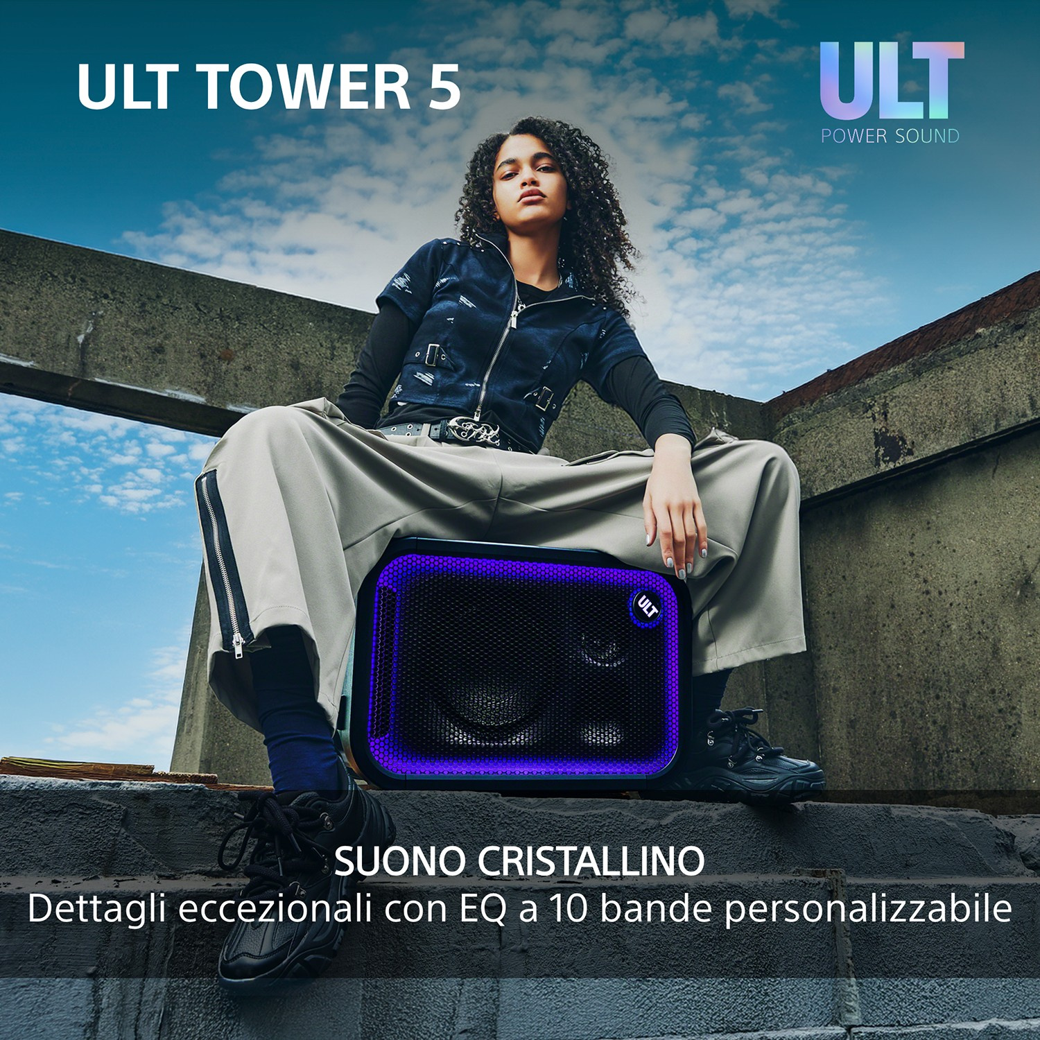 Una donna siede su un grande speaker ULT TOWER 5 all'aperto, con uno sfondo di cielo azzurro nuvoloso.