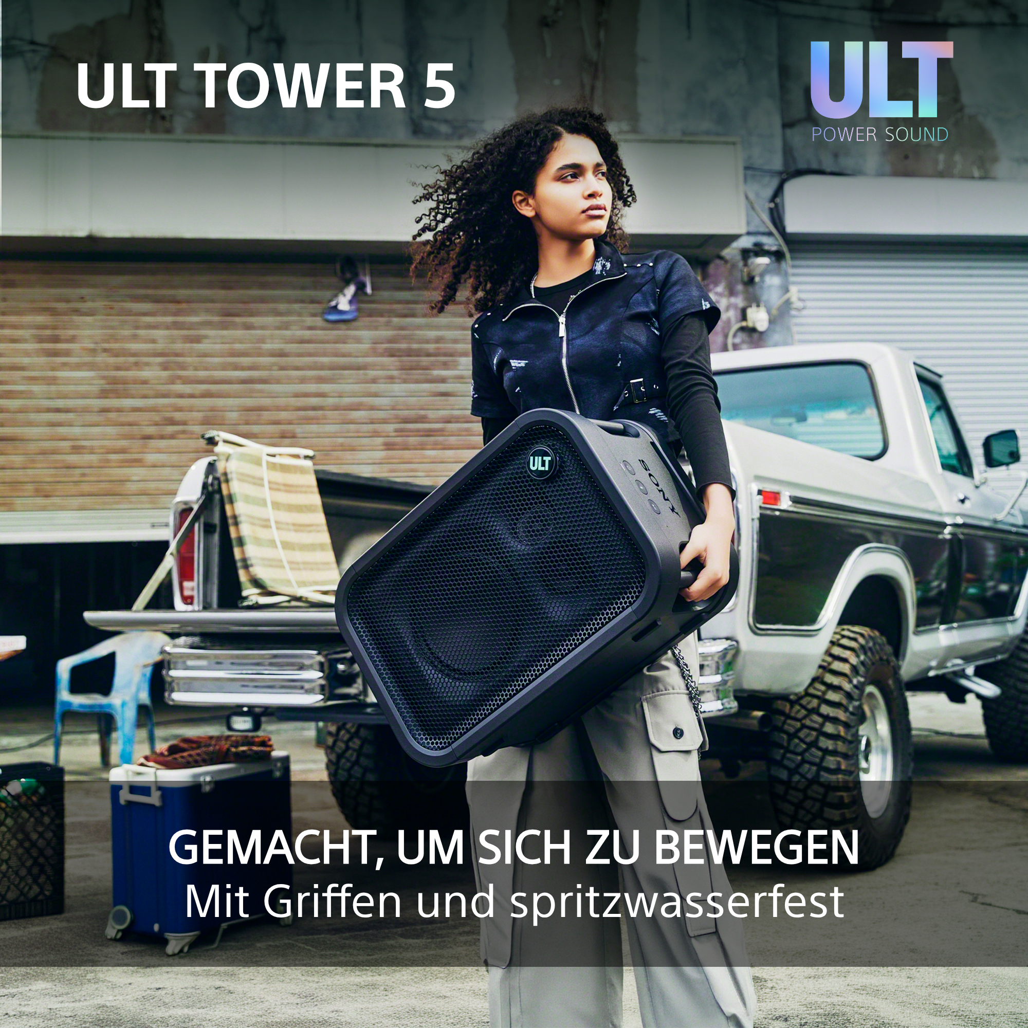 Eine Person mit lockigem Haar trägt einen großen schwarzen Lautsprecher vor einem weißen Pick-up.