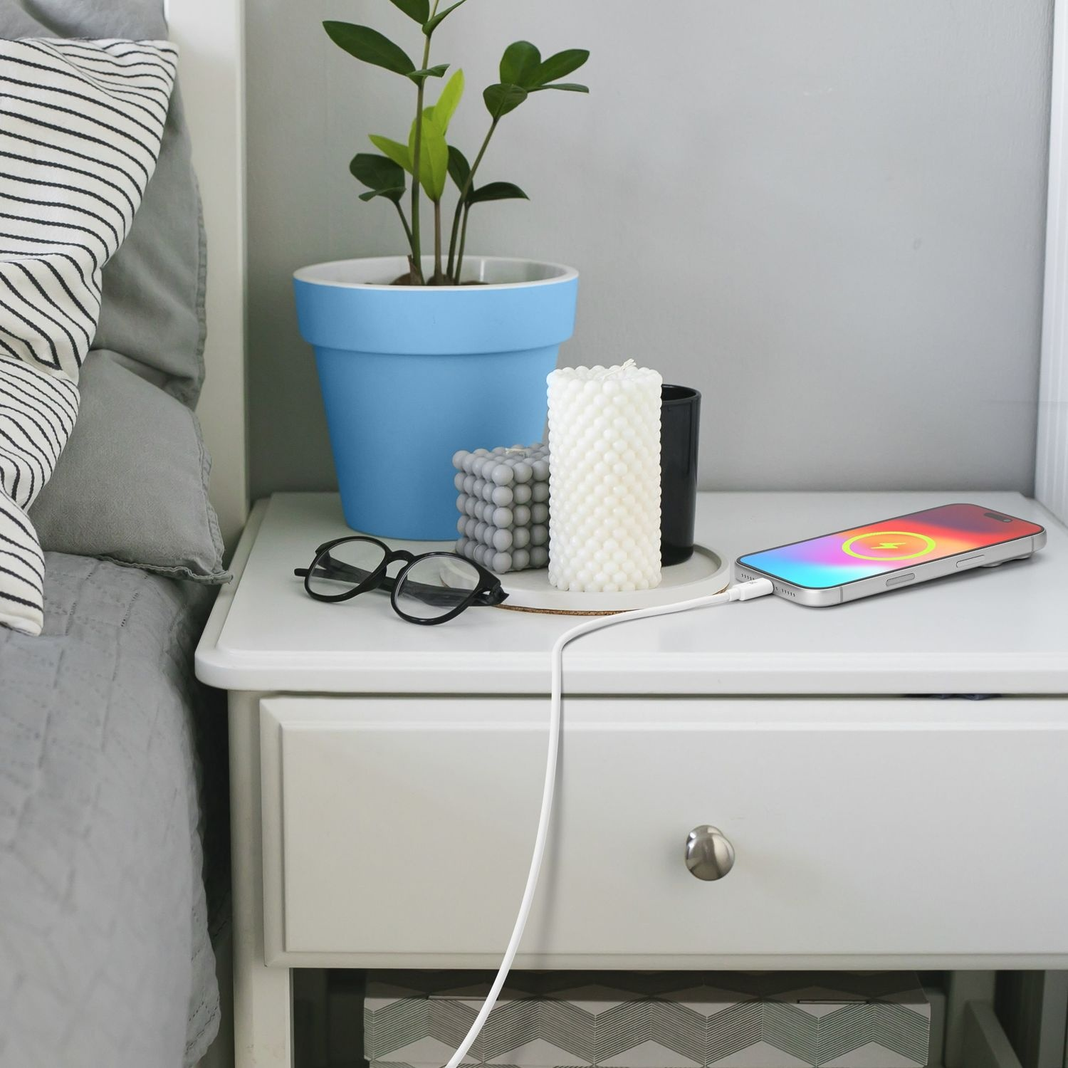 Un comodino bianco con occhiali, candele, un vaso blu e uno smartphone in carica accanto a un letto grigio.