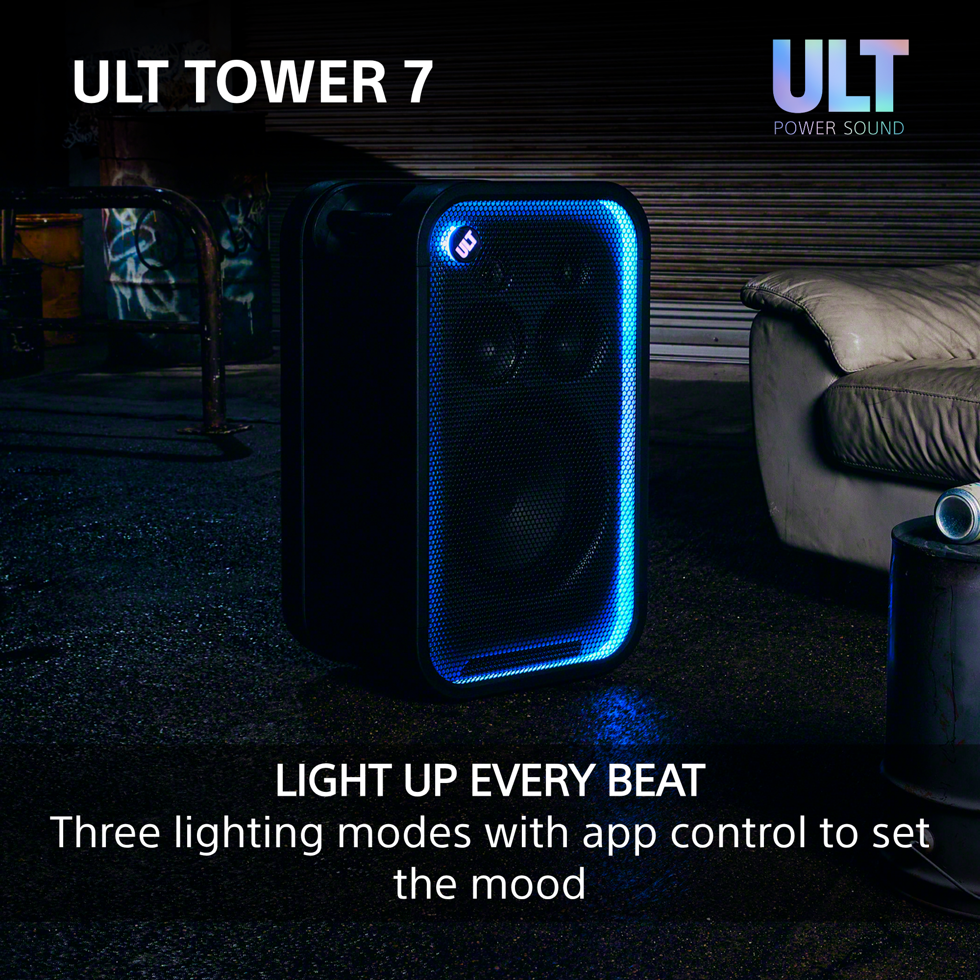 Schwarzer ULT Tower 7 Lautsprecher mit blauer Randbeleuchtung in einem Raum mit Couch und Metallfass.