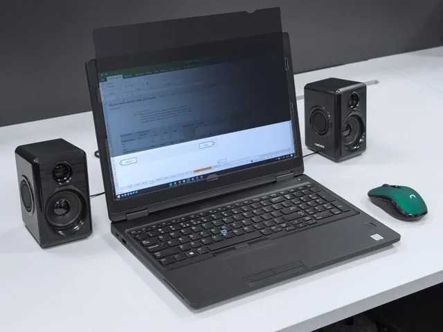 Czarny laptop z filtrem prywatności, dwoma głośnikami i zieloną myszą na białym biurku.
