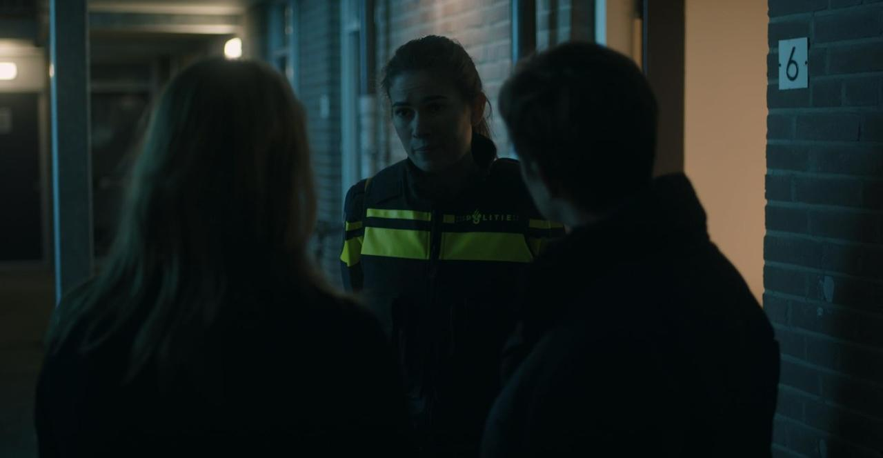 Eine Polizistin in Uniform spricht in einem Flur vor einer Tür mit der Nummer sechs mit zwei Personen.