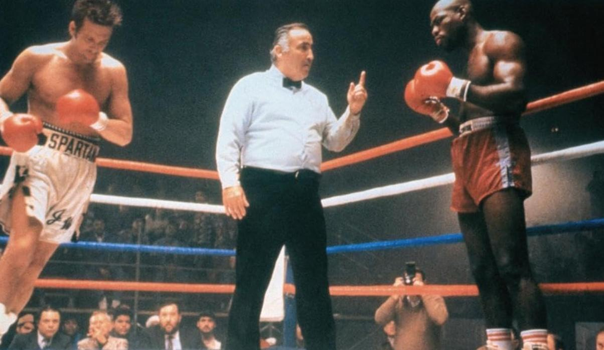Ein Schiedsrichter in weißem Hemd und schwarzer Hose steht zwischen zwei Boxern im Ring.