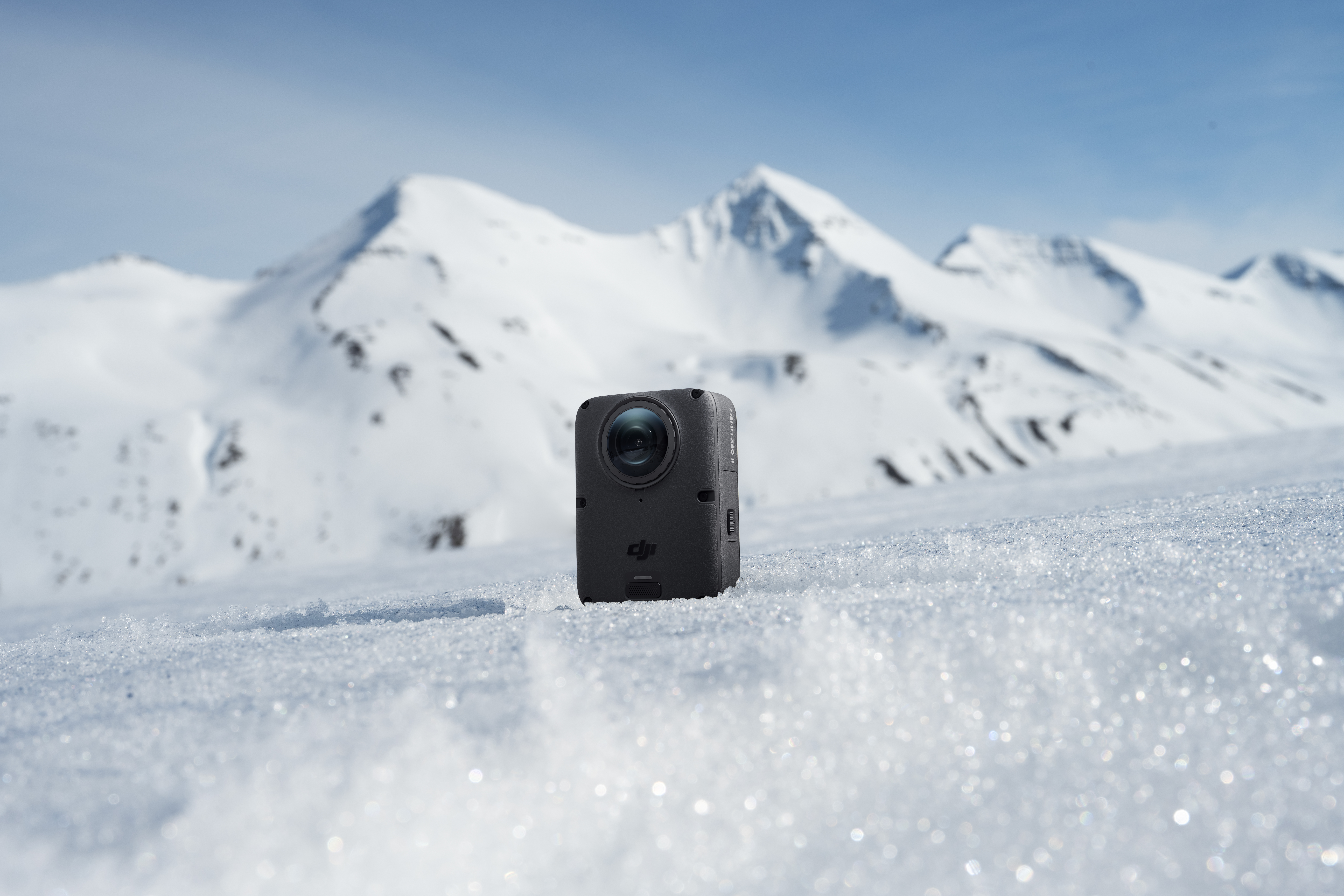 Eine schwarze DJI-Kamera steht im Schnee vor einer verschneiten Bergkette unter blauem Himmel.