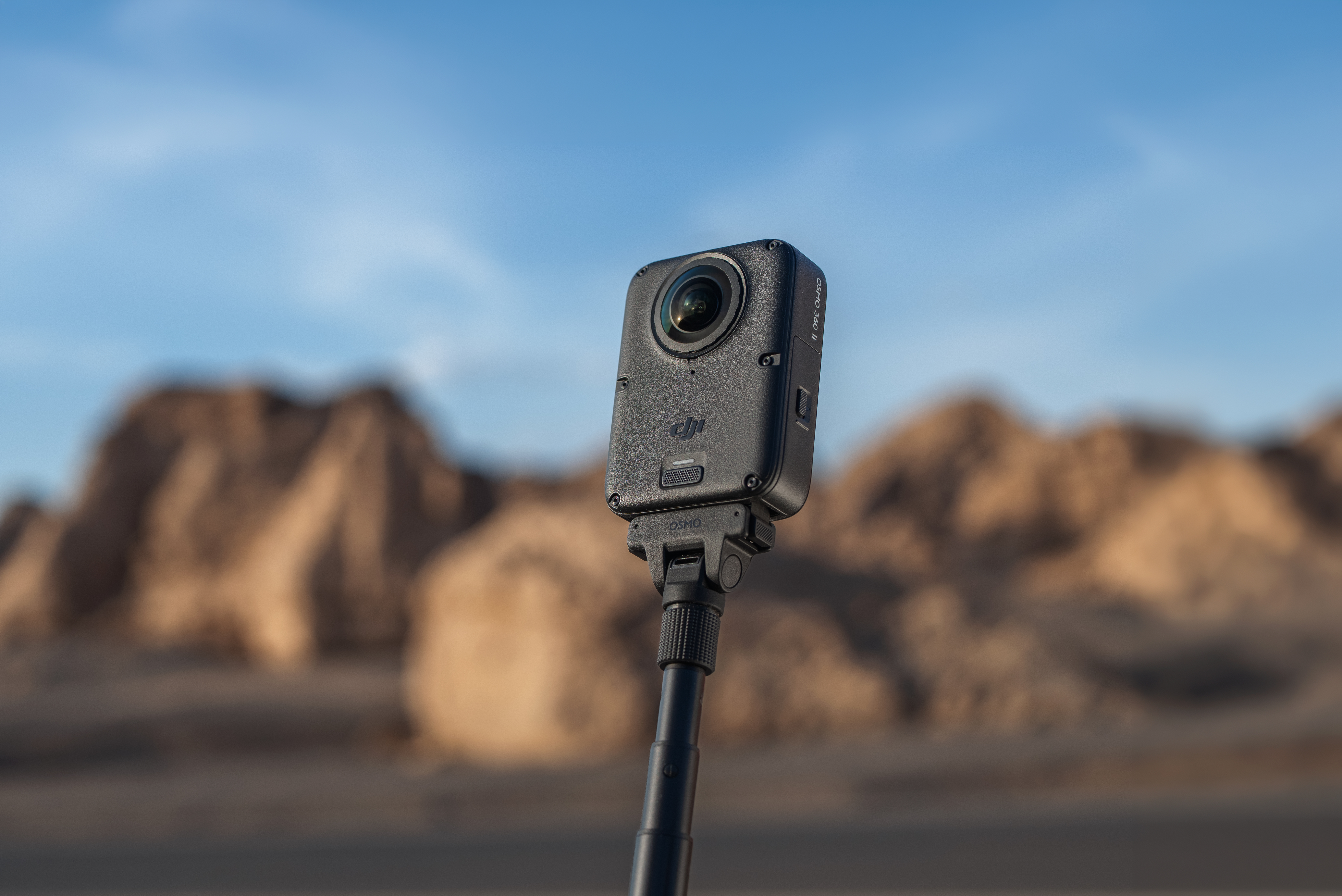 Eine schwarze DJI Osmo Action-Kamera auf einem Selfie-Stick vor einer unscharfen Felslandschaft unter blauem Himmel.