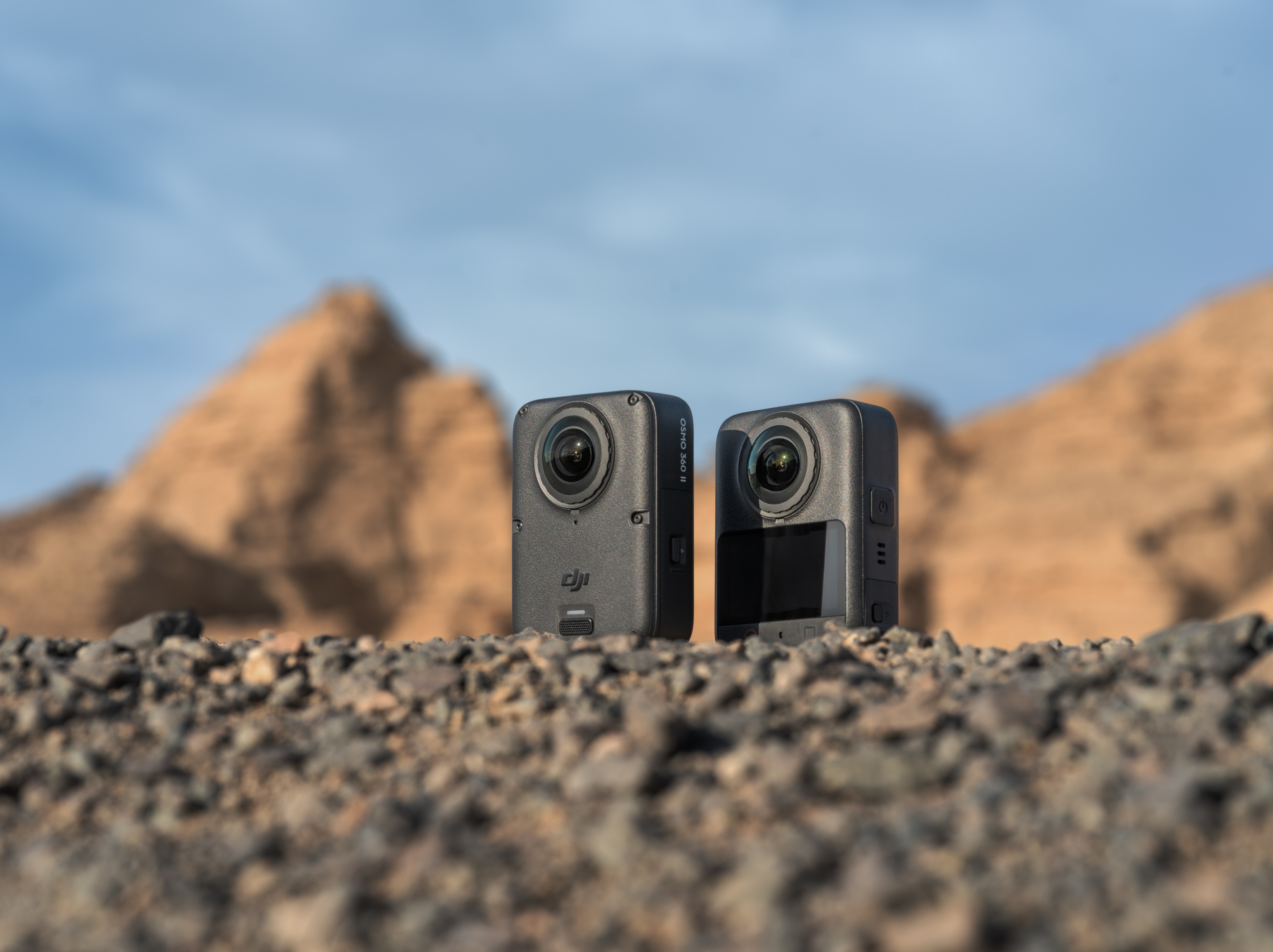 Zwei Osmo 360-Kameras auf steinigem Boden mit Bergen und blauem Himmel im Hintergrund.