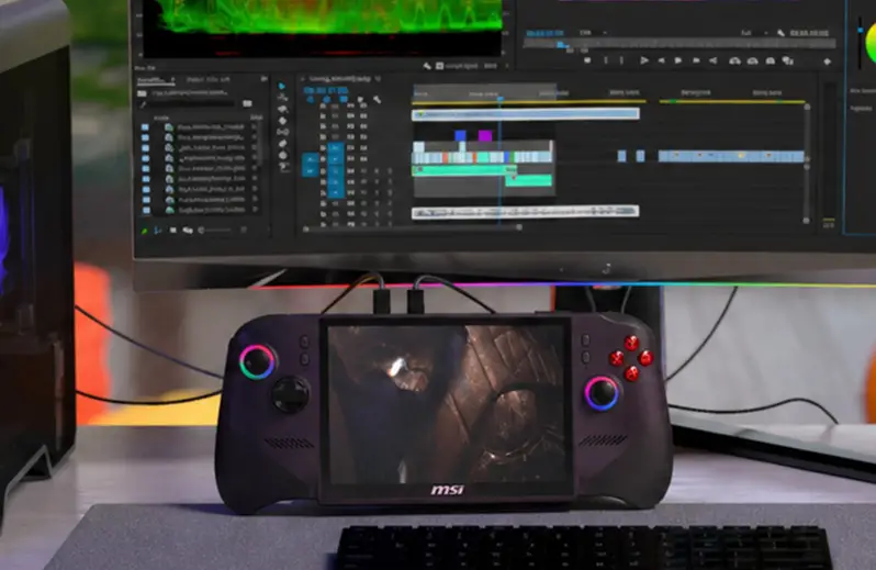 Una console di gioco portatile con anelli luminosi sui joystick è posizionata su una scrivania davanti a un grande monitor.