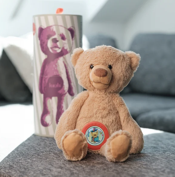 Ein Plüschbär sitzt auf einer grauen Couch neben einer zylindrischen Verpackung mit einem Bärenmotiv.