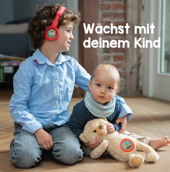 Ein Junge mit roten Kopfhörern sitzt neben einem Baby mit einem Plüschtier auf dem Holzfussboden.